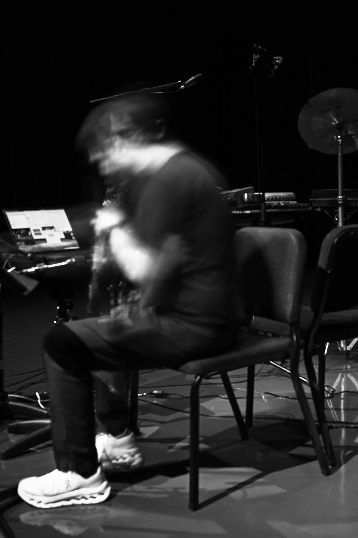 A person sitting on a chair in a dark room, surrounded by musical equipment, with a laptop open in front of them, and blurred motion suggesting movement.