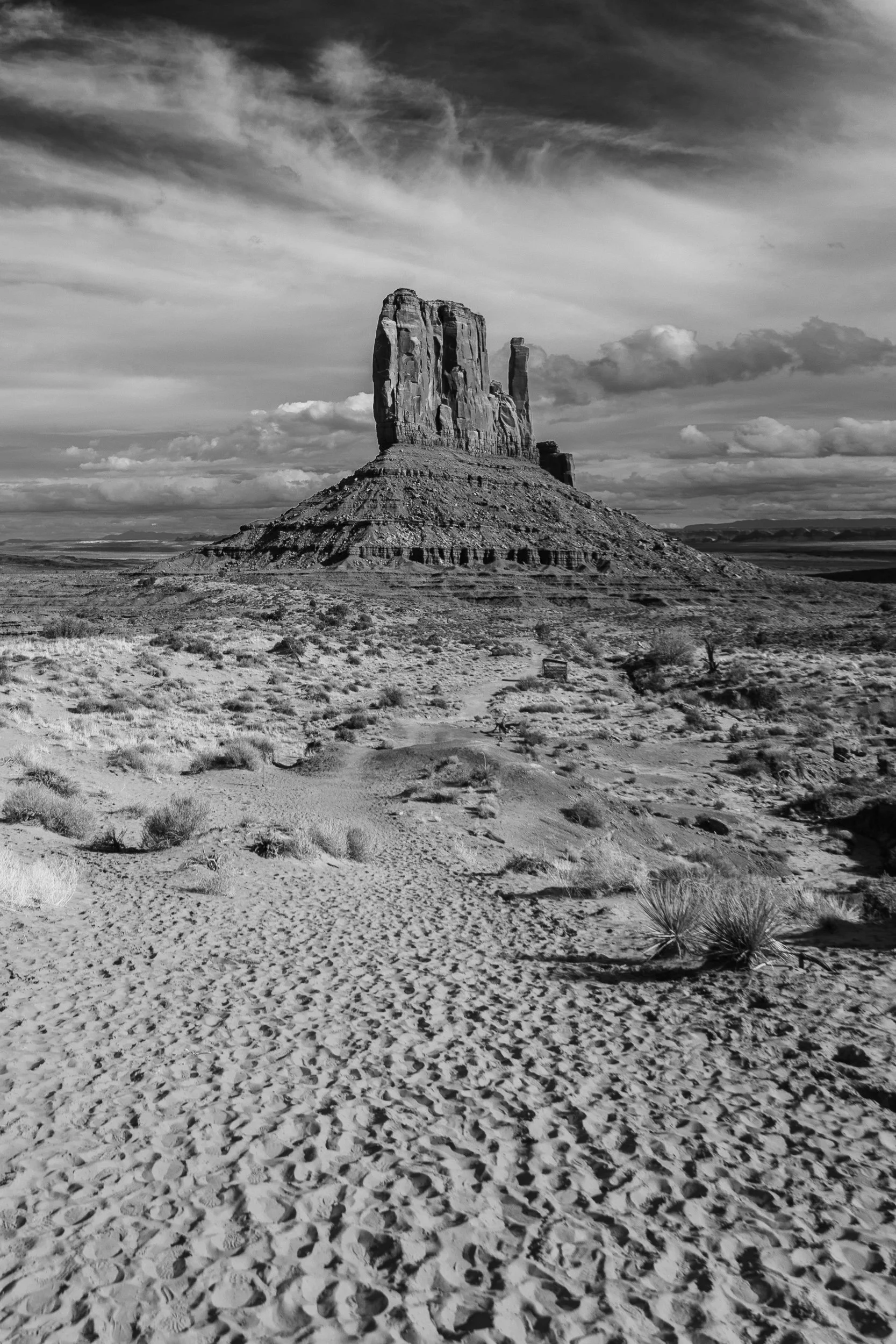 Black and white photo of a large rock formation with a flat top in a desert landscape, with scattered bushes and a sky filled with clouds.