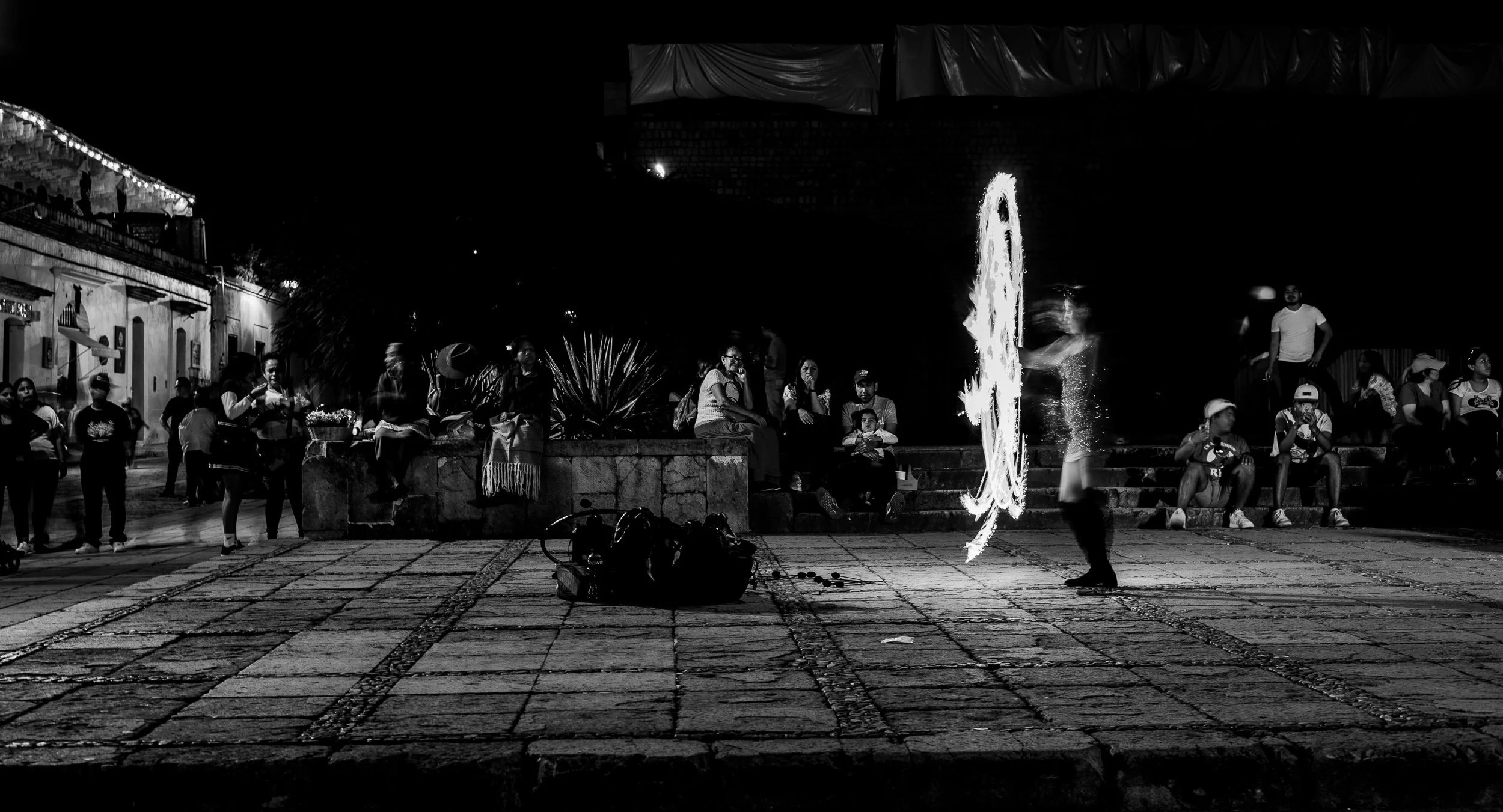 A street performer in front of a crowd at night, creating fire patterns with a flaming tool. People are sitting and standing around, watching the performance. Some individuals have cameras or phones, capturing the moment. The scene is in black and wh