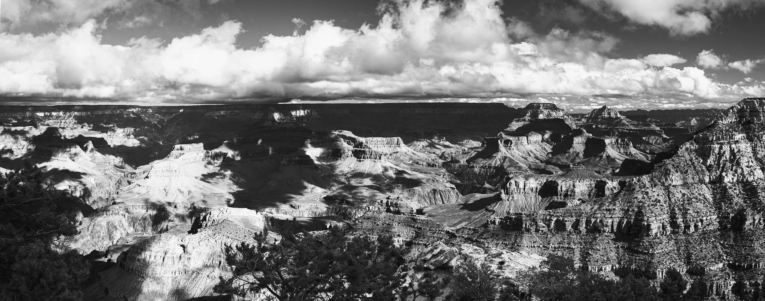 Black and white photograph of the Grand Canyon with layered rock formations and clouds overhead.