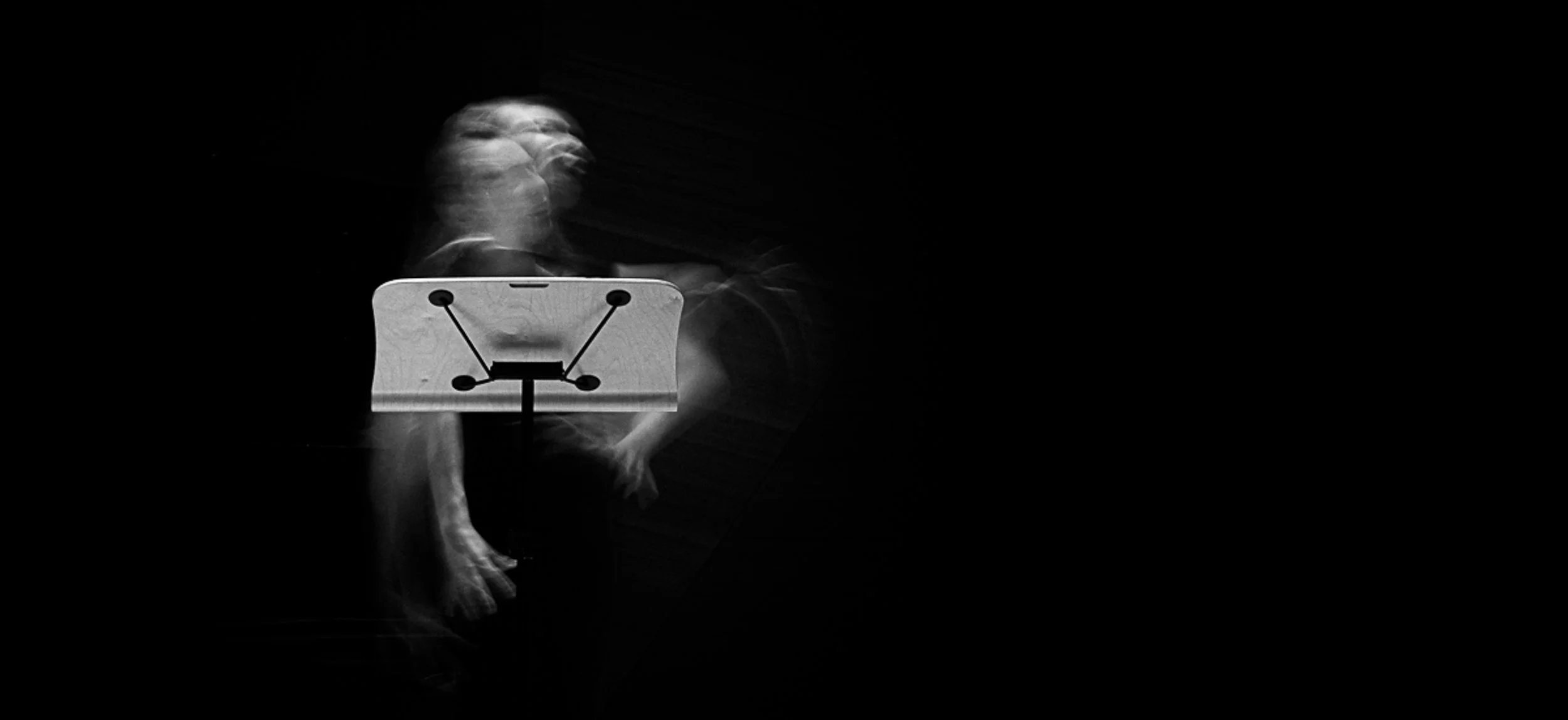 Blurred image of a person standing behind a music stand, with movement causing a ghostly double exposure effect, set against a black background.