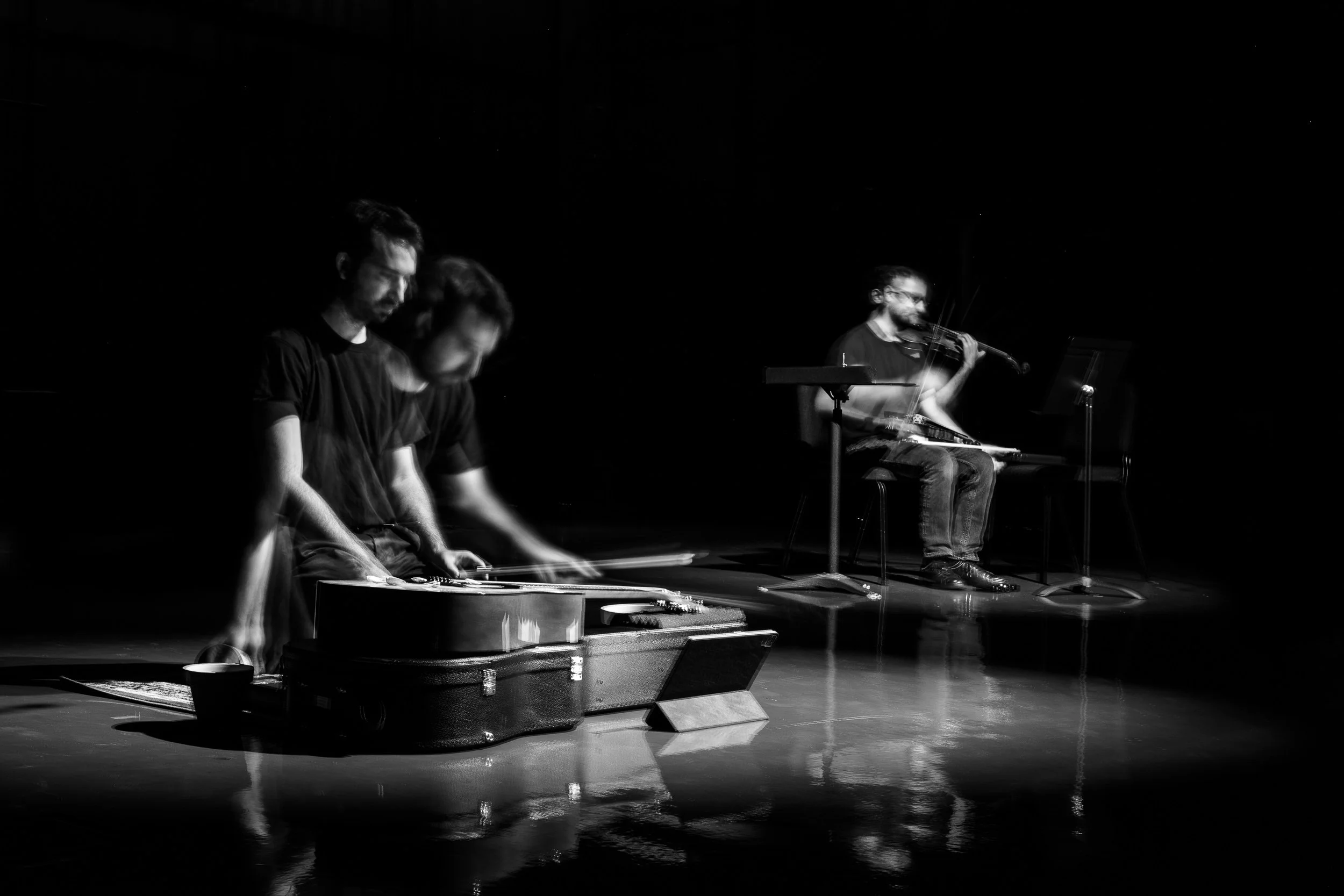 Two musicians playing instruments on a dark stage, one with a guitar and another with a violin, with motion blur indicating movement.