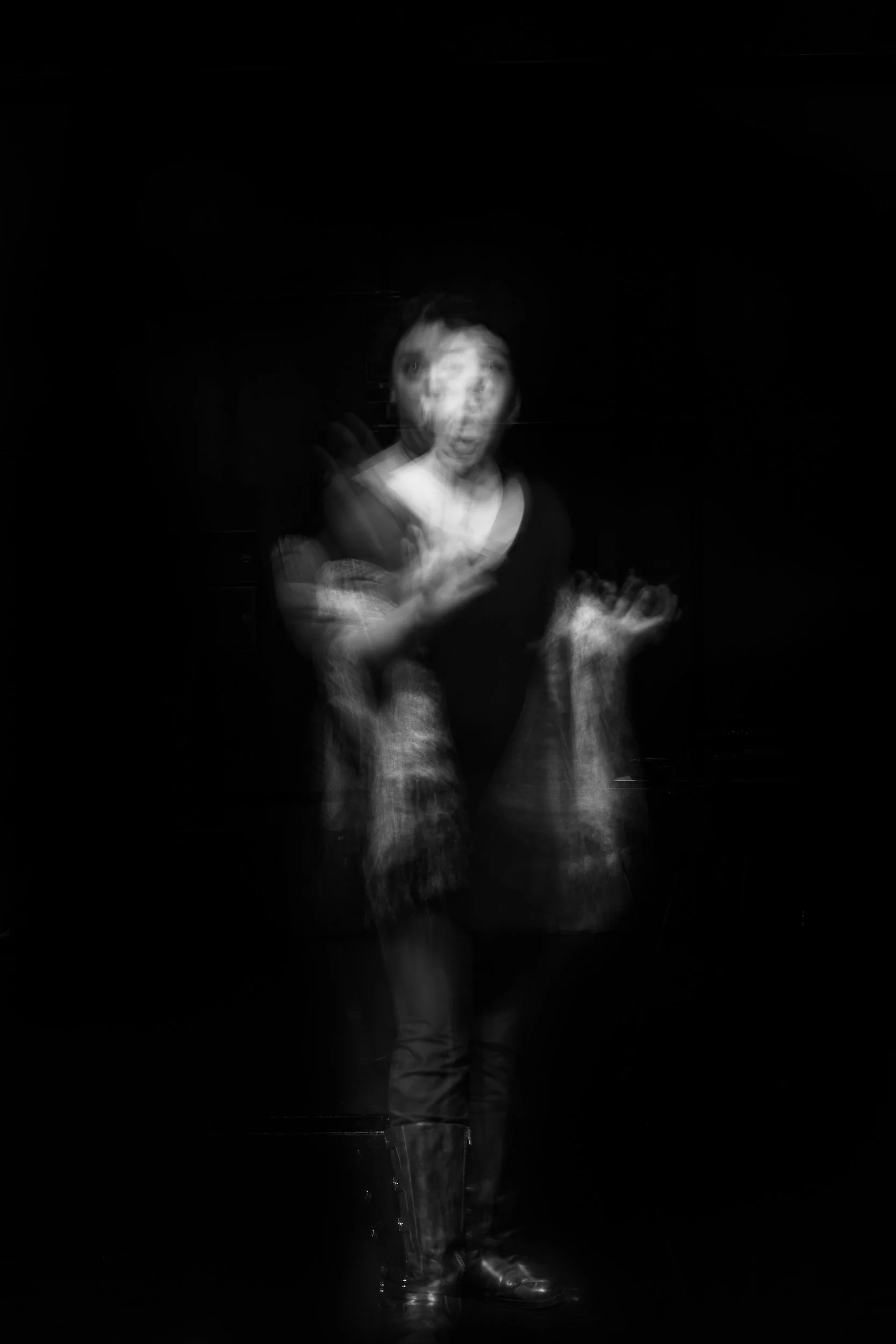 Blurry black and white photo of a woman with short hair, wearing a black top, jeans, and boots, standing against a dark background, with motion blur and double exposure effects.