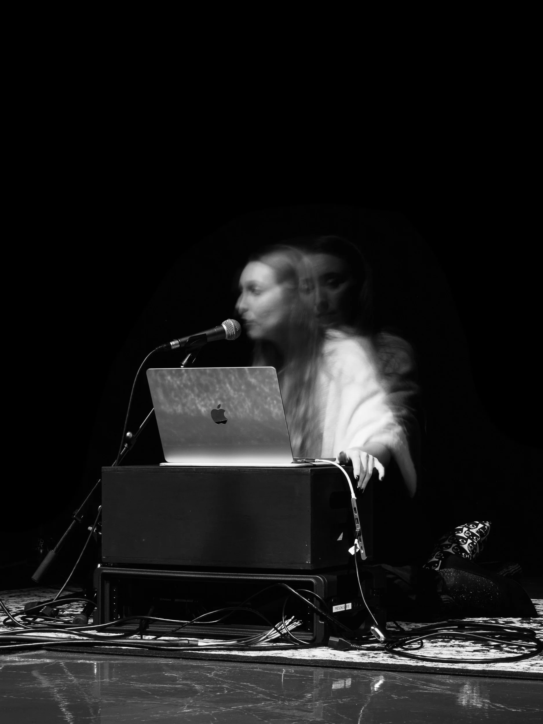 A woman with long hair and light-colored clothing is performing on stage with a microphone, a laptop on a stand, and various cables and equipment on the floor. The image has a motion blur effect.