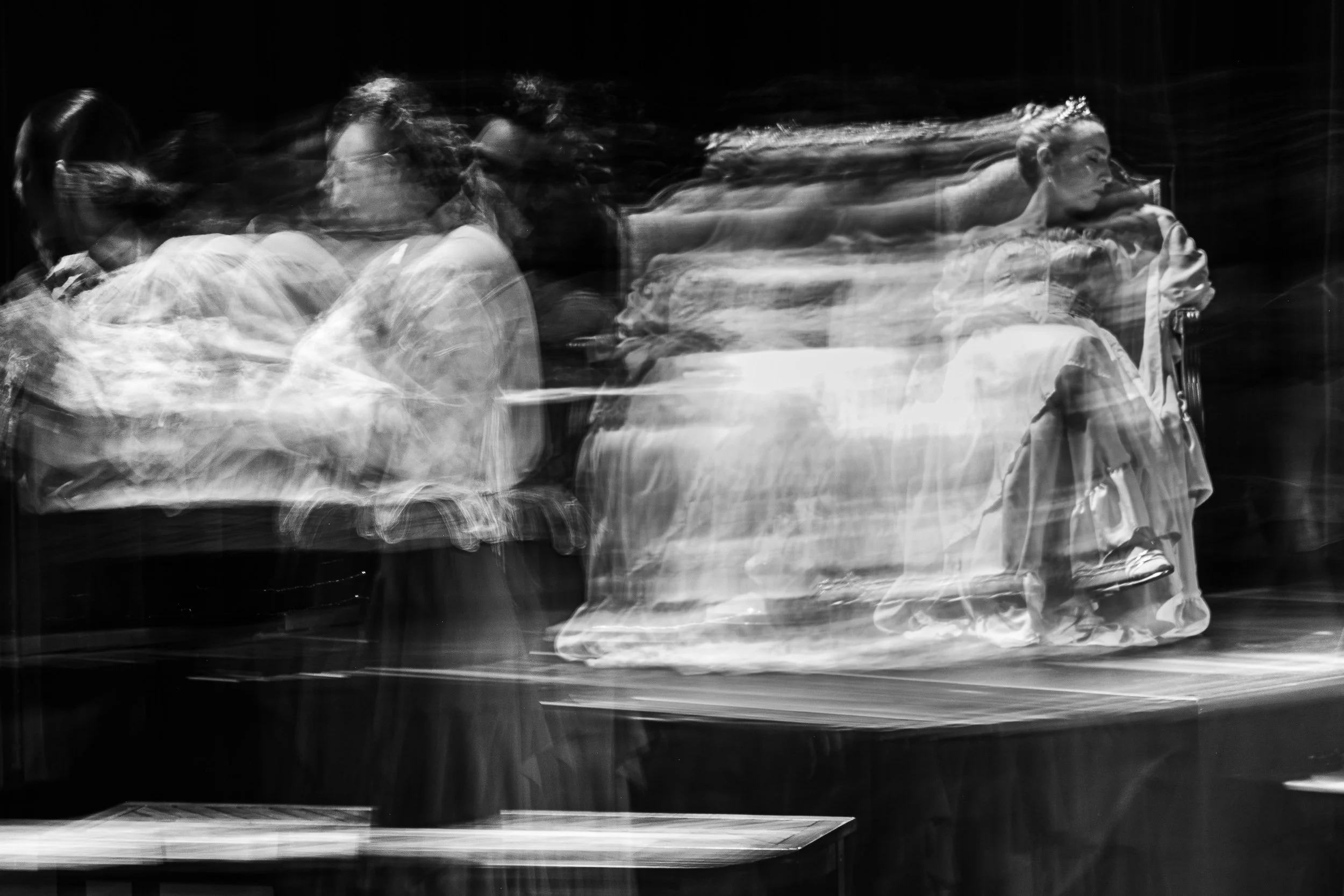 Black and white photo of a woman sitting on a chair, with a motion blur effect creating multiple overlapping images of her in different positions.