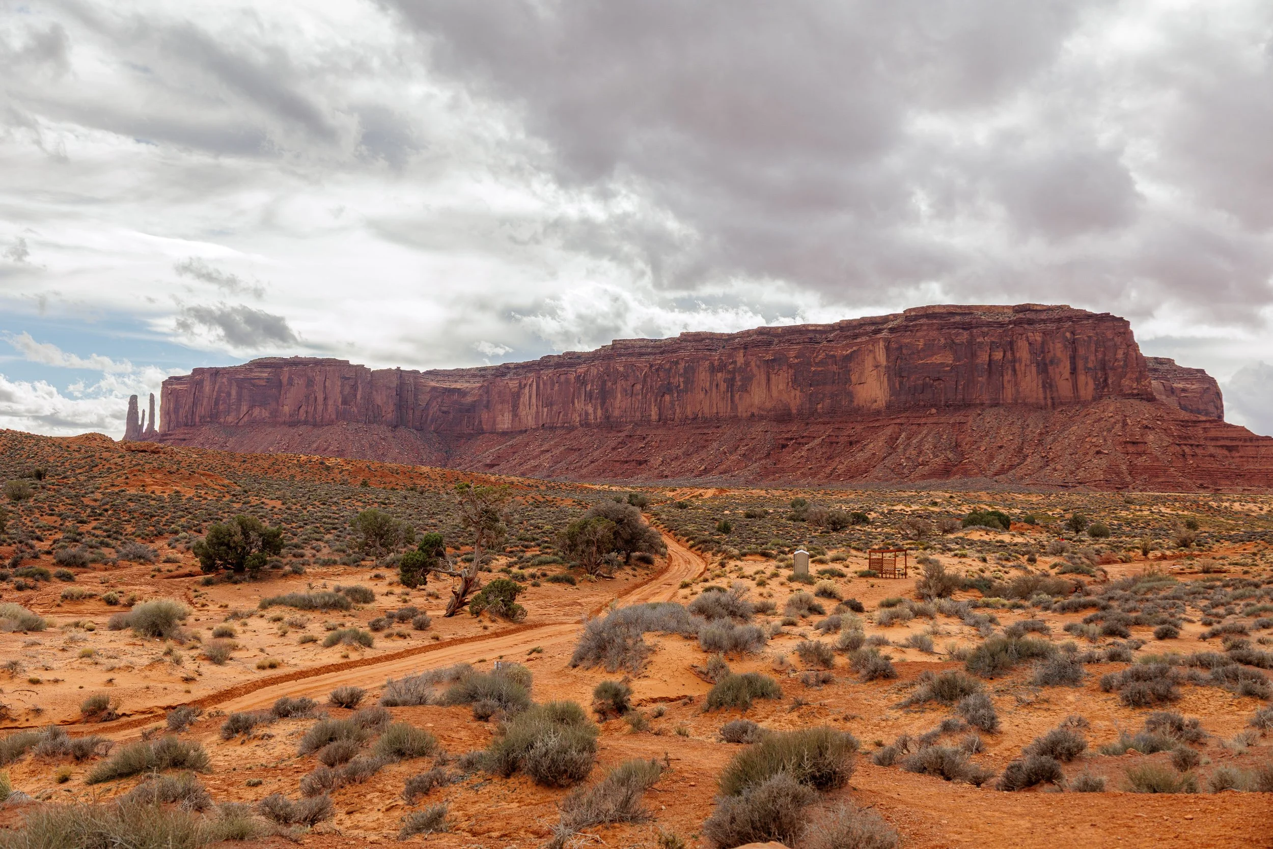 A desert landscape with a large red rock formation under cloudy skies, sparse vegetation, and a dirt path winding through the terrain.