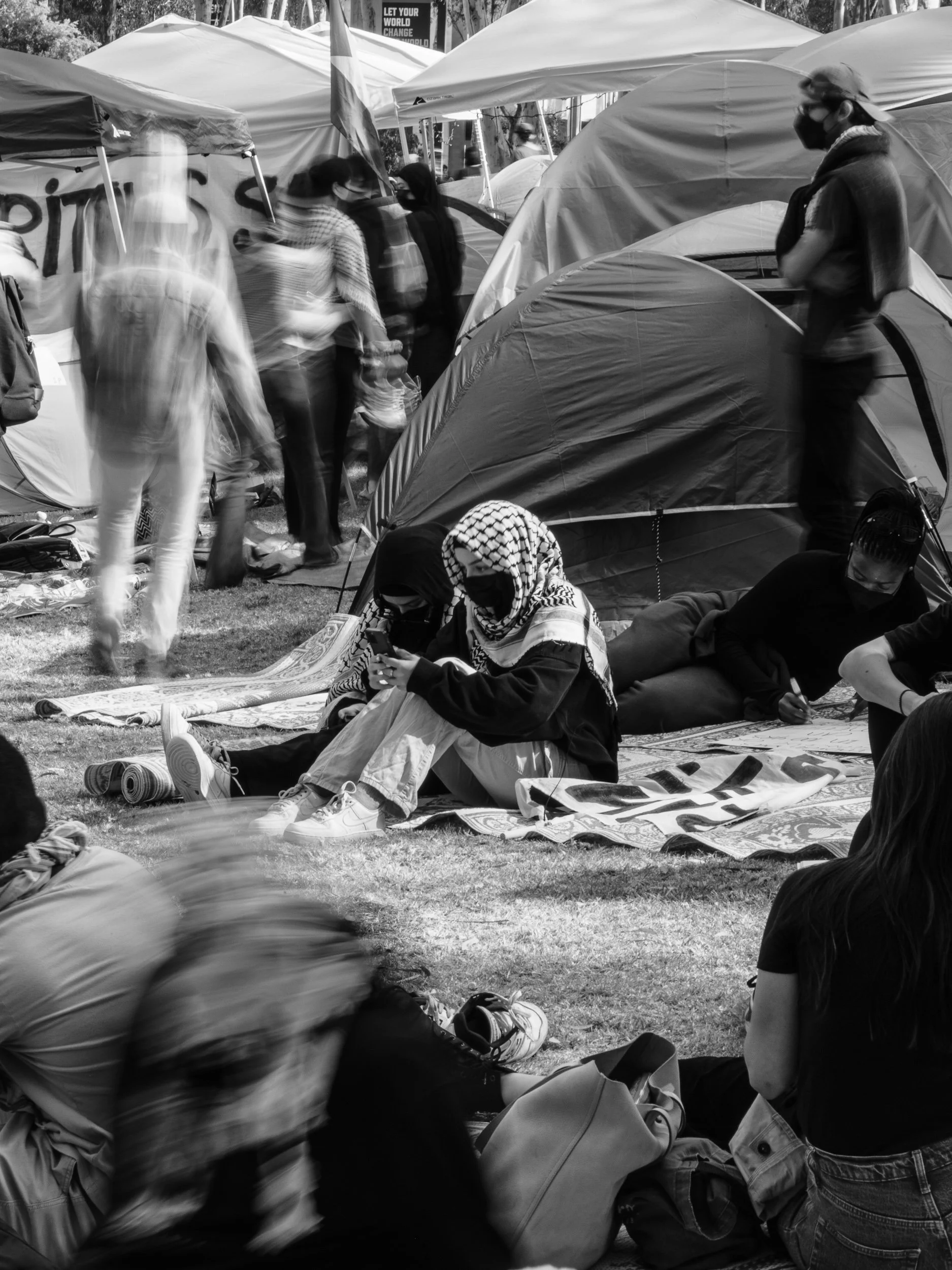 People sitting and standing among tents at a protest or outdoor gathering, some wearing masks and headscarves, with blurred motion of other attendees in the background.