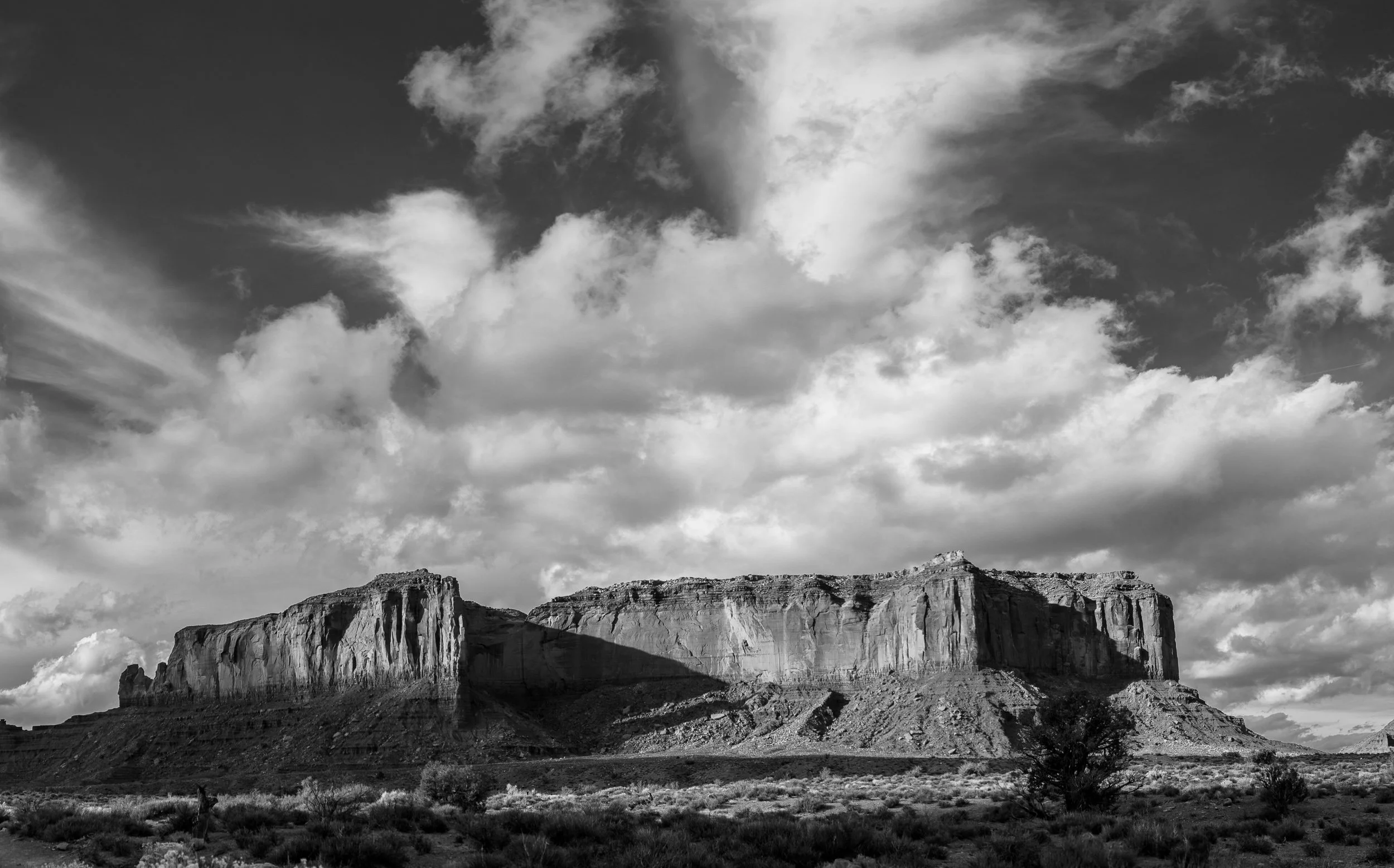 Black and white photo of large flat-topped mesas in a desert landscape with dramatic clouds in the sky.