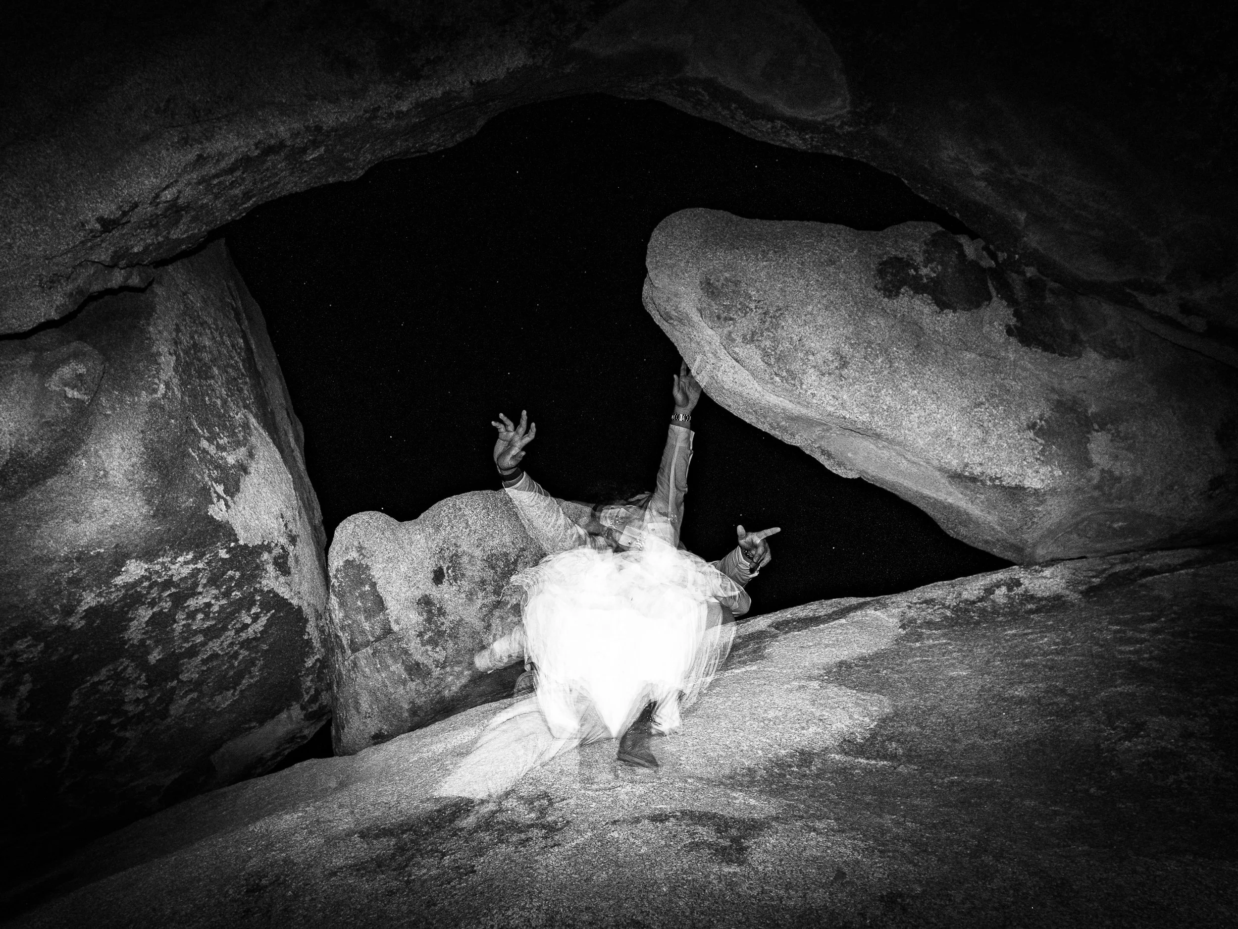 A person lying on rocky ground in a cave, with arms raised and pointing, black and white photo.