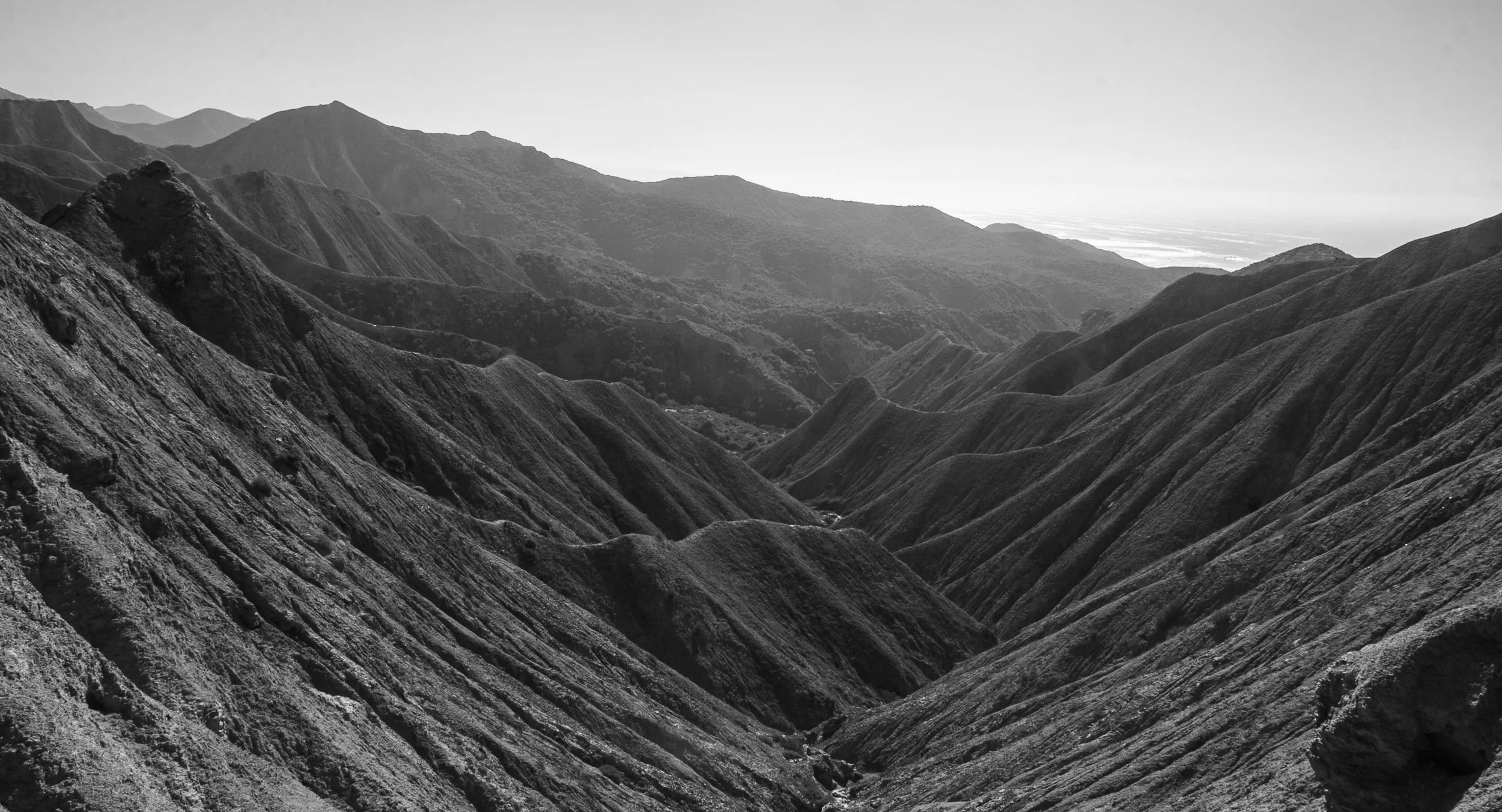 Black and white photograph of rugged mountain ridges with deep valleys and rolling hills in the distance.