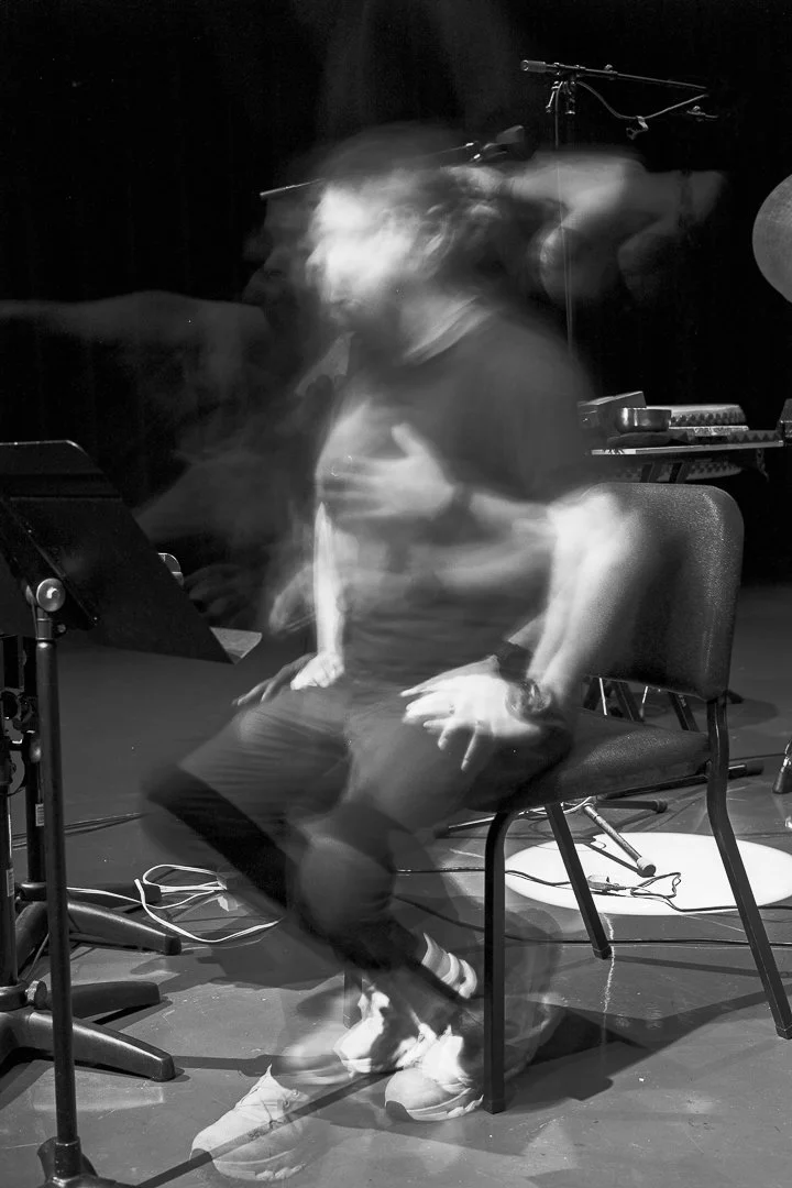 A person sitting on a chair surrounded by musical equipment, captured with motion blur, in black and white.