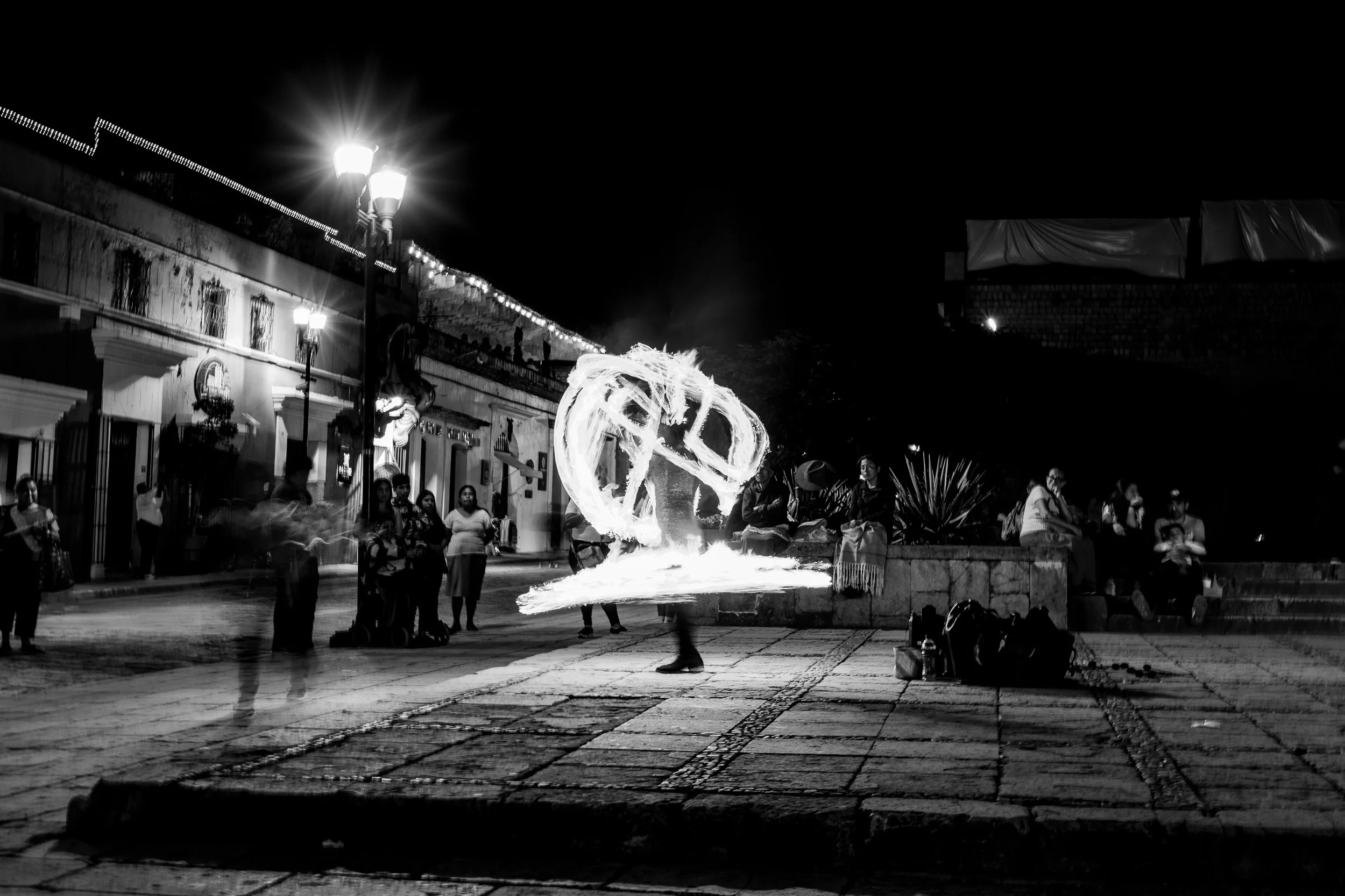Night scene of a street performance with a fire dancer creating light patterns with fire, surrounded by an audience on a city street.