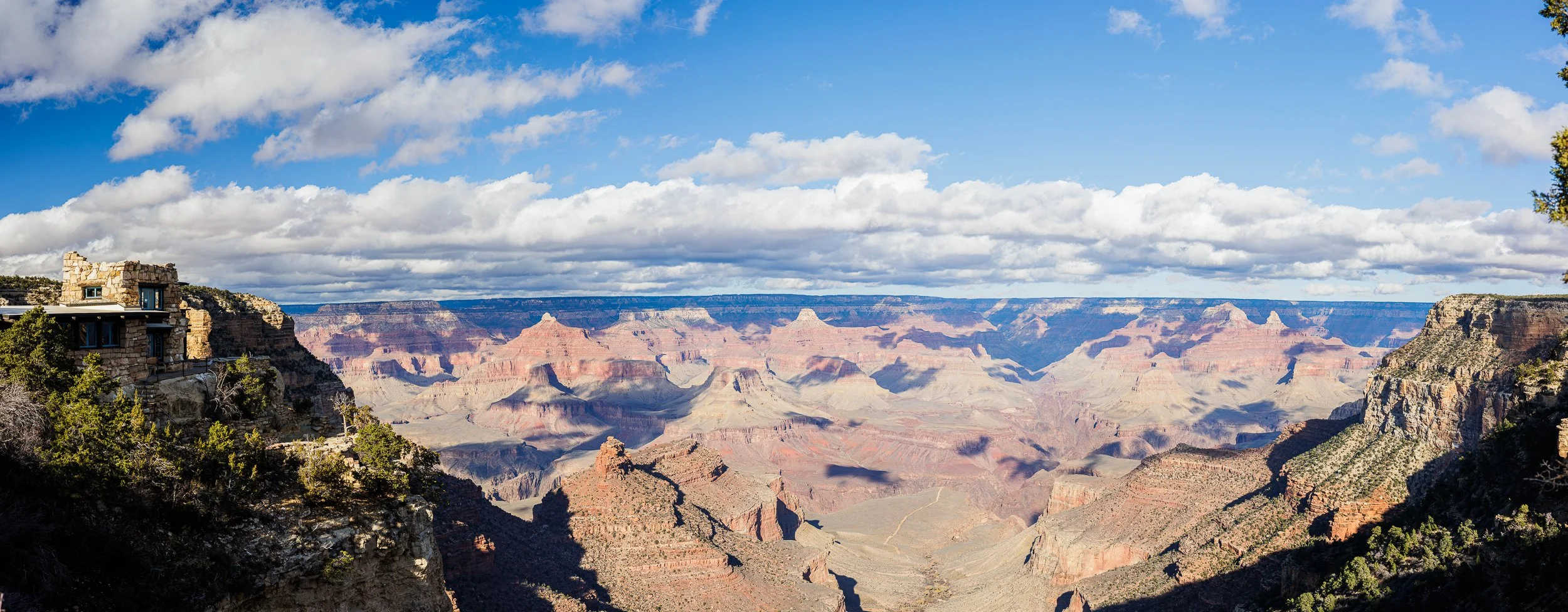 View of the Grand Canyon under a partly cloudy sky with a building on the left on a rocky ledge.