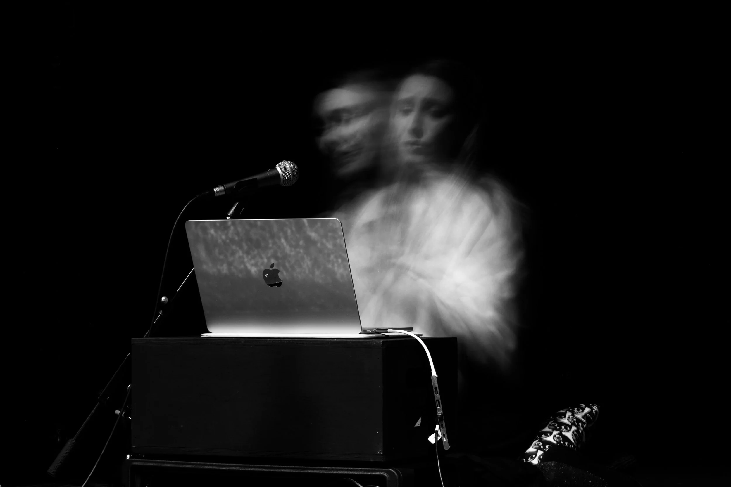 A person at a desk with a laptop and microphone, appearing to be in motion, creating a blurred, ghostly effect, against a dark background.