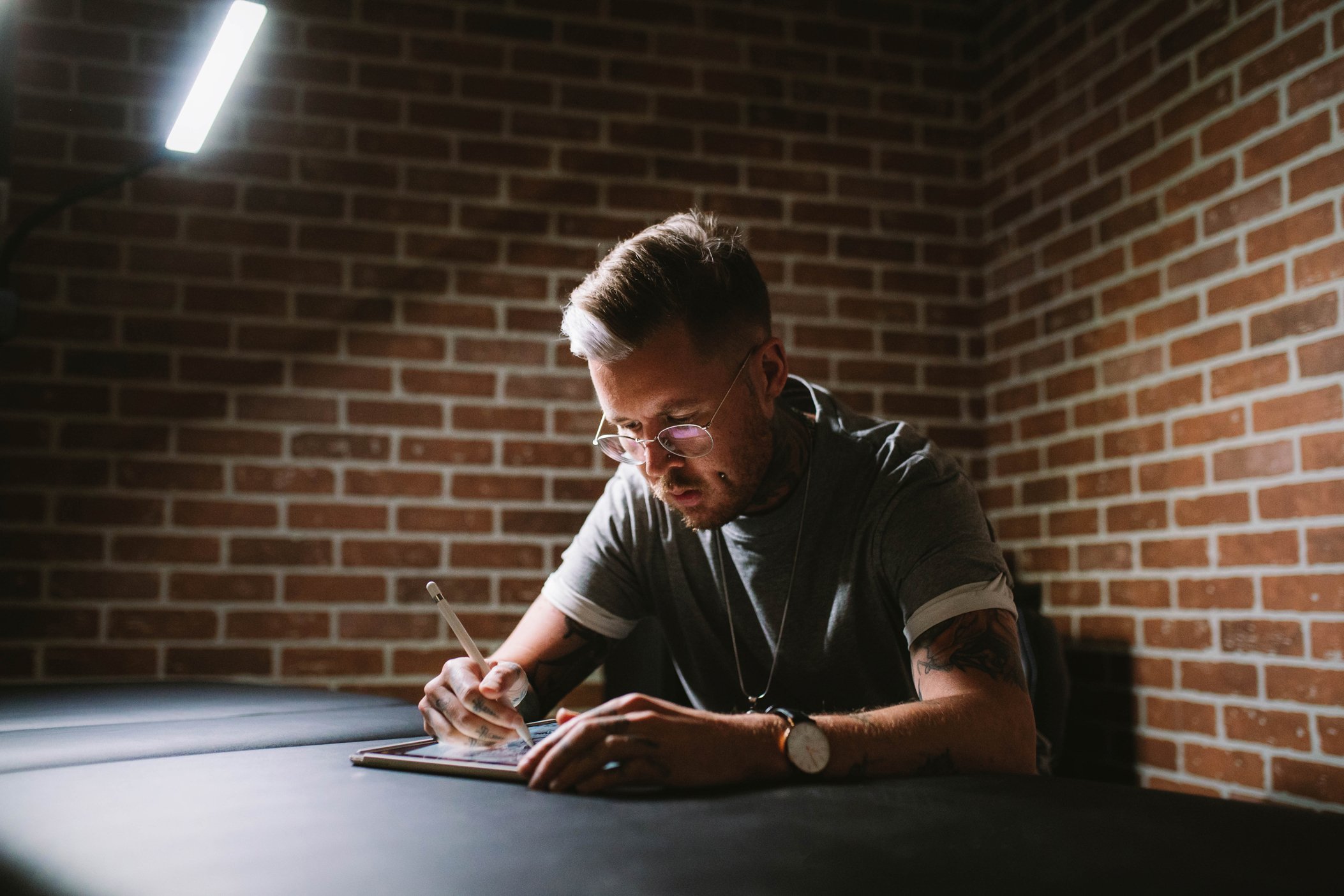 Young man with glasses and tattoos writing in a notebook at a desk in a dimly lit room with brick wall background.