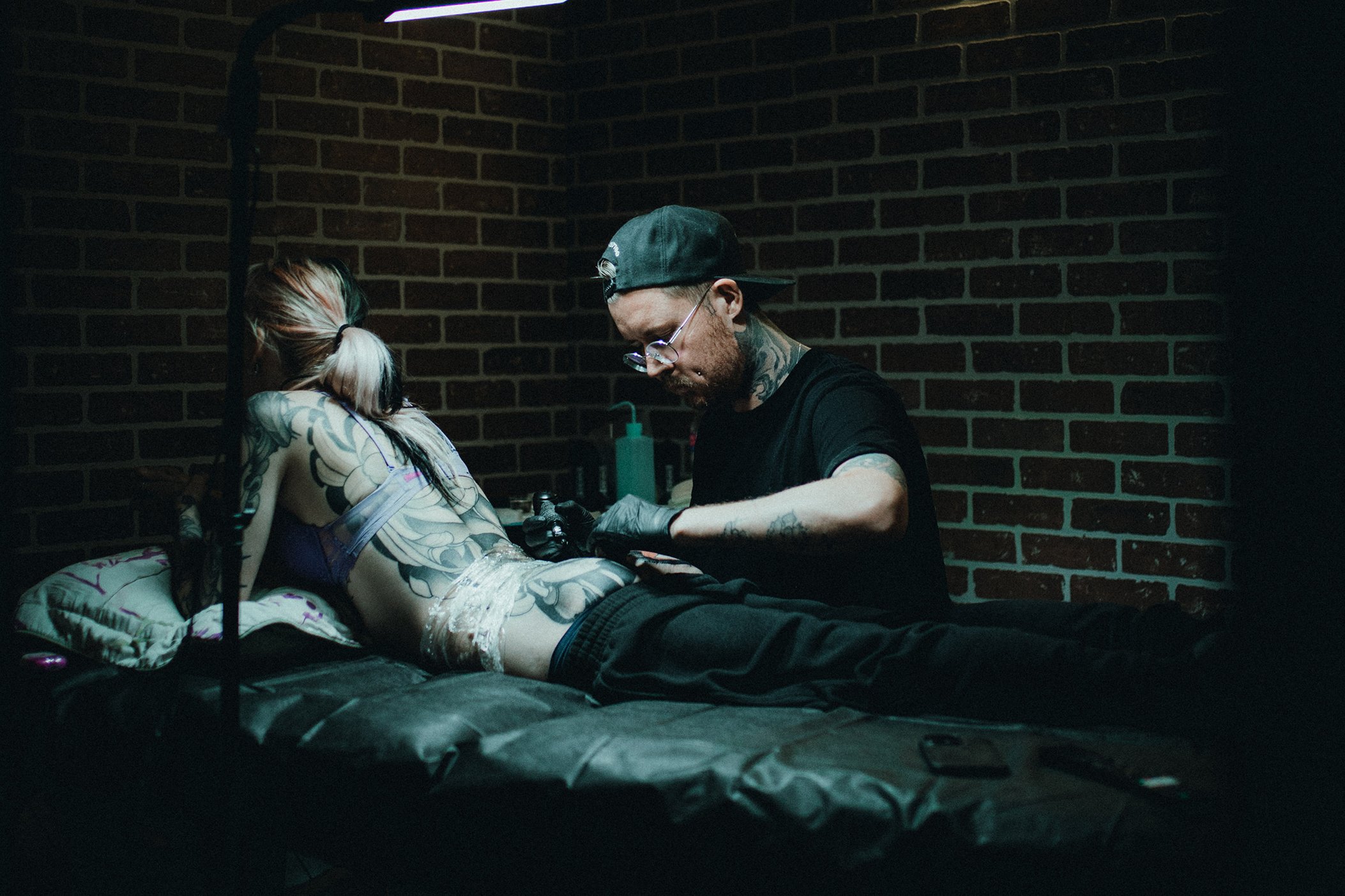 Tattoo artist working on a client's back in a tattoo studio with brick wall background.