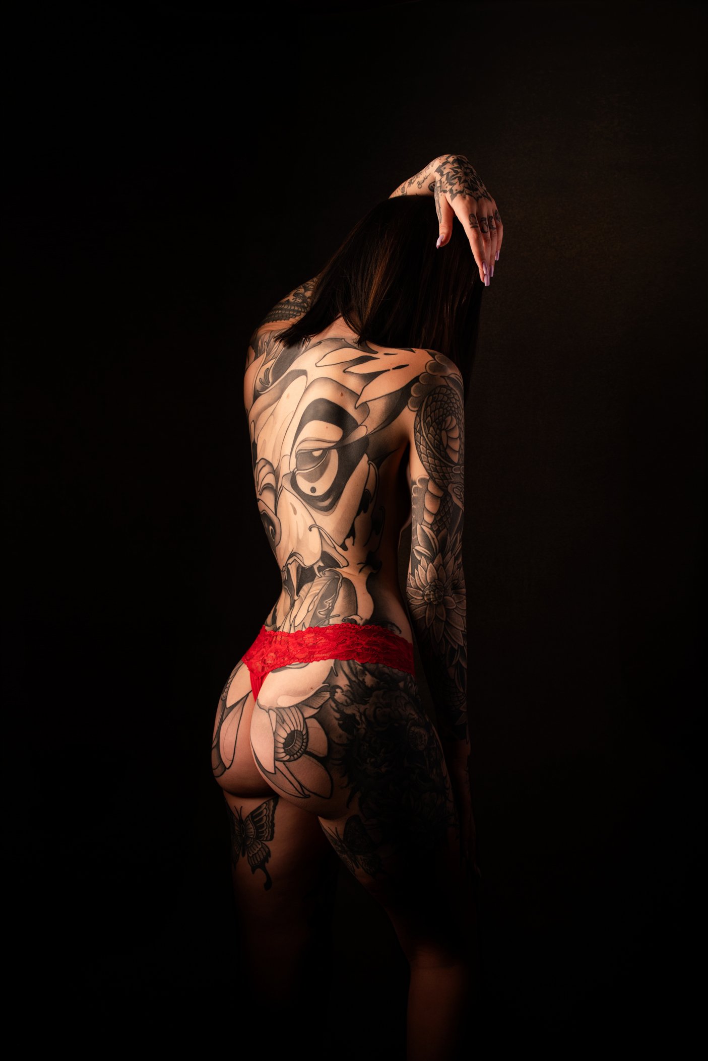 A woman with numerous black tattoos covering her back, arms, and legs, wearing red lace underwear, standing against a black background with her head bowed and her left hand resting on her head.