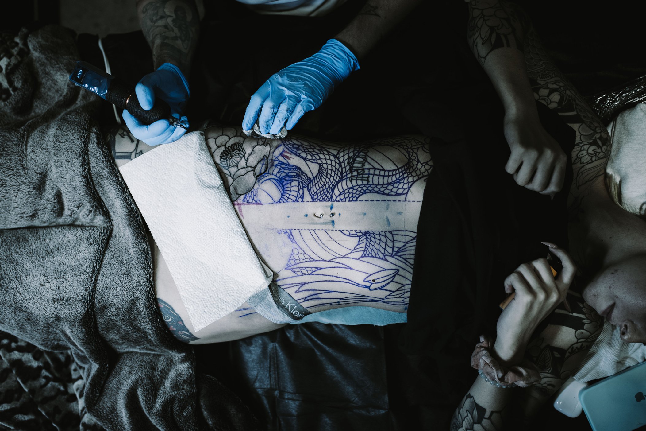 A woman lying on a table undergoing a tattoo session, with a tattoo artist using equipment to create a tattoo on her abdomen. The woman has multiple tattoos, and the artist is wearing blue gloves.