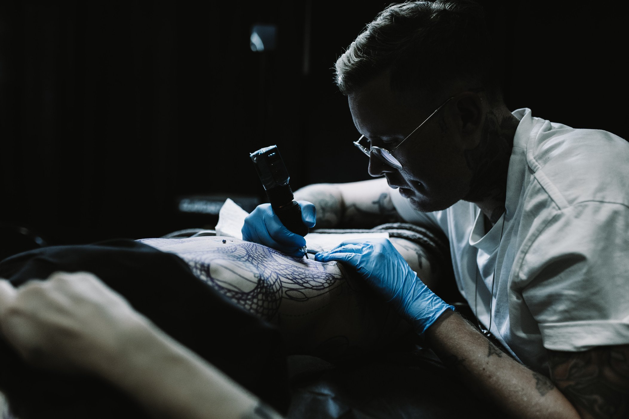 A tattoo artist wearing glasses and blue gloves creating a tattoo on a person's arm with a tattoo machine in a dark studio.