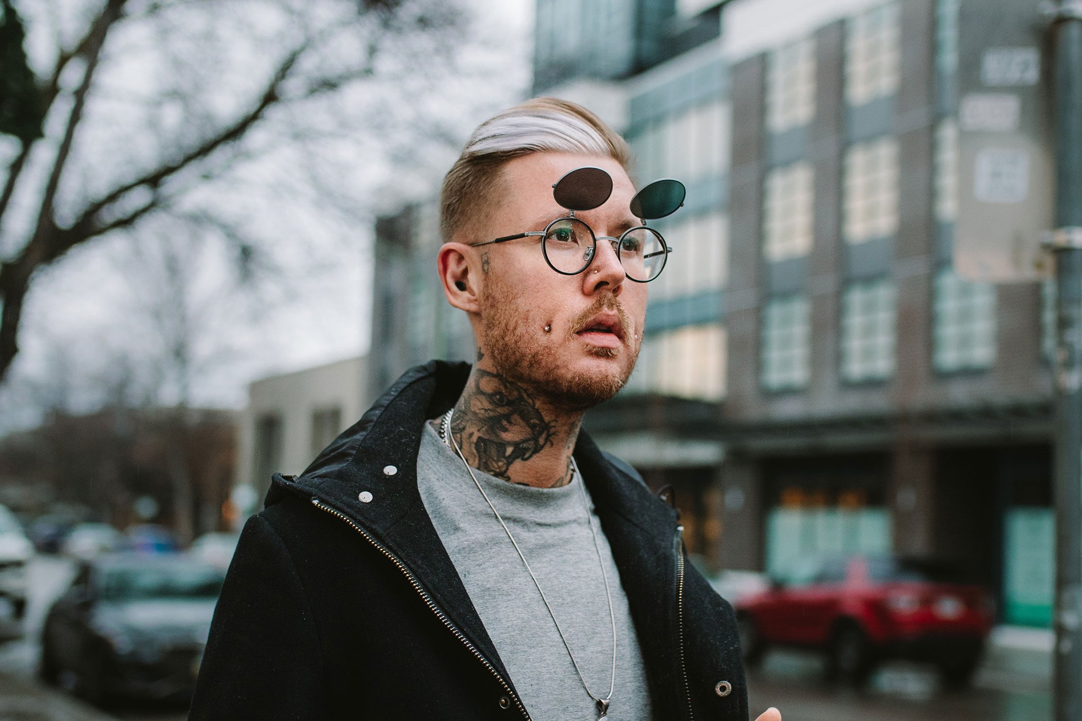 A young man with beige hair, glasses, tattoos, and facial piercings standing outdoors on a city street