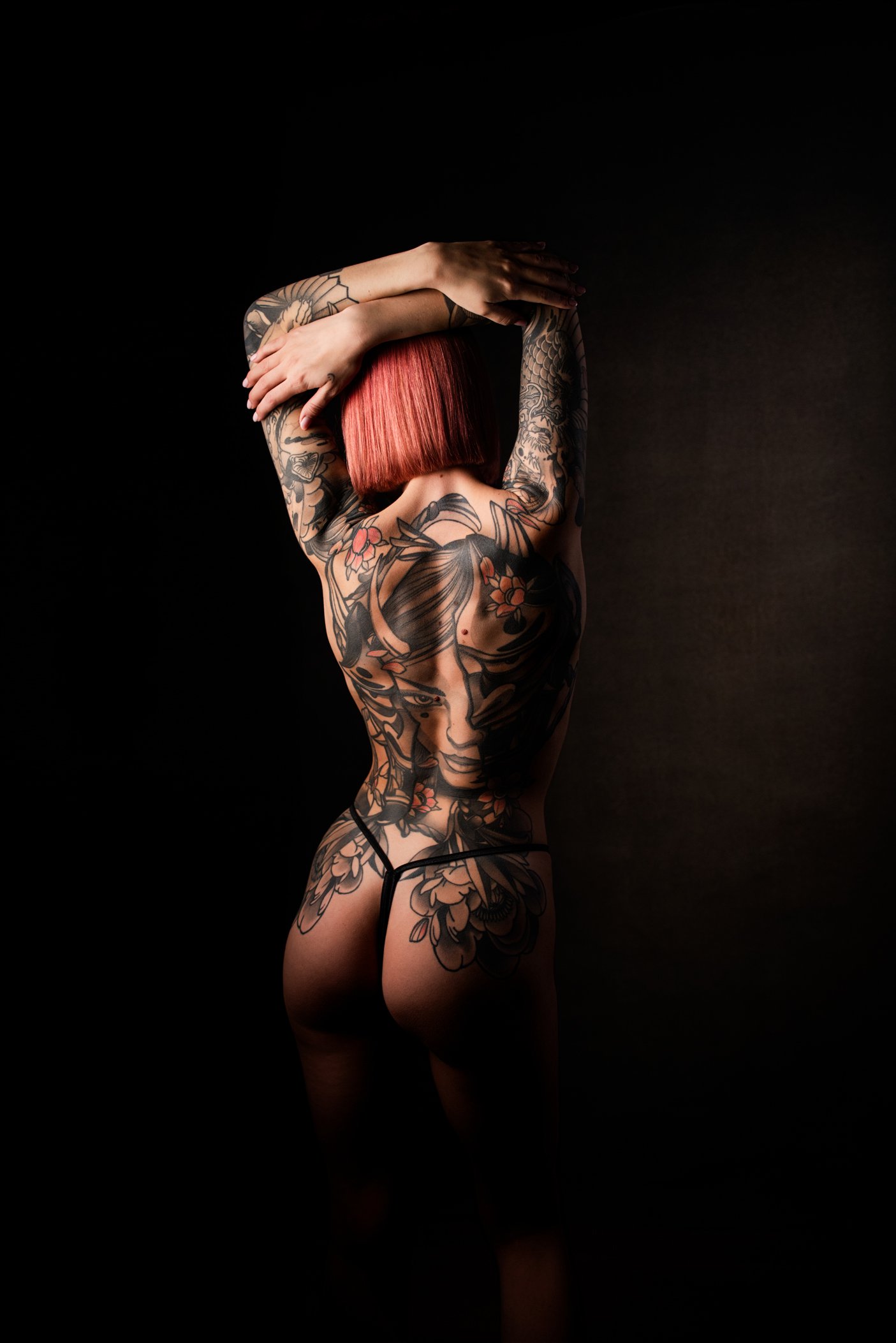 A woman with red hair and extensive tattoos on her back, arms, and legs, standing against a dark background with her arms raised above her head.
