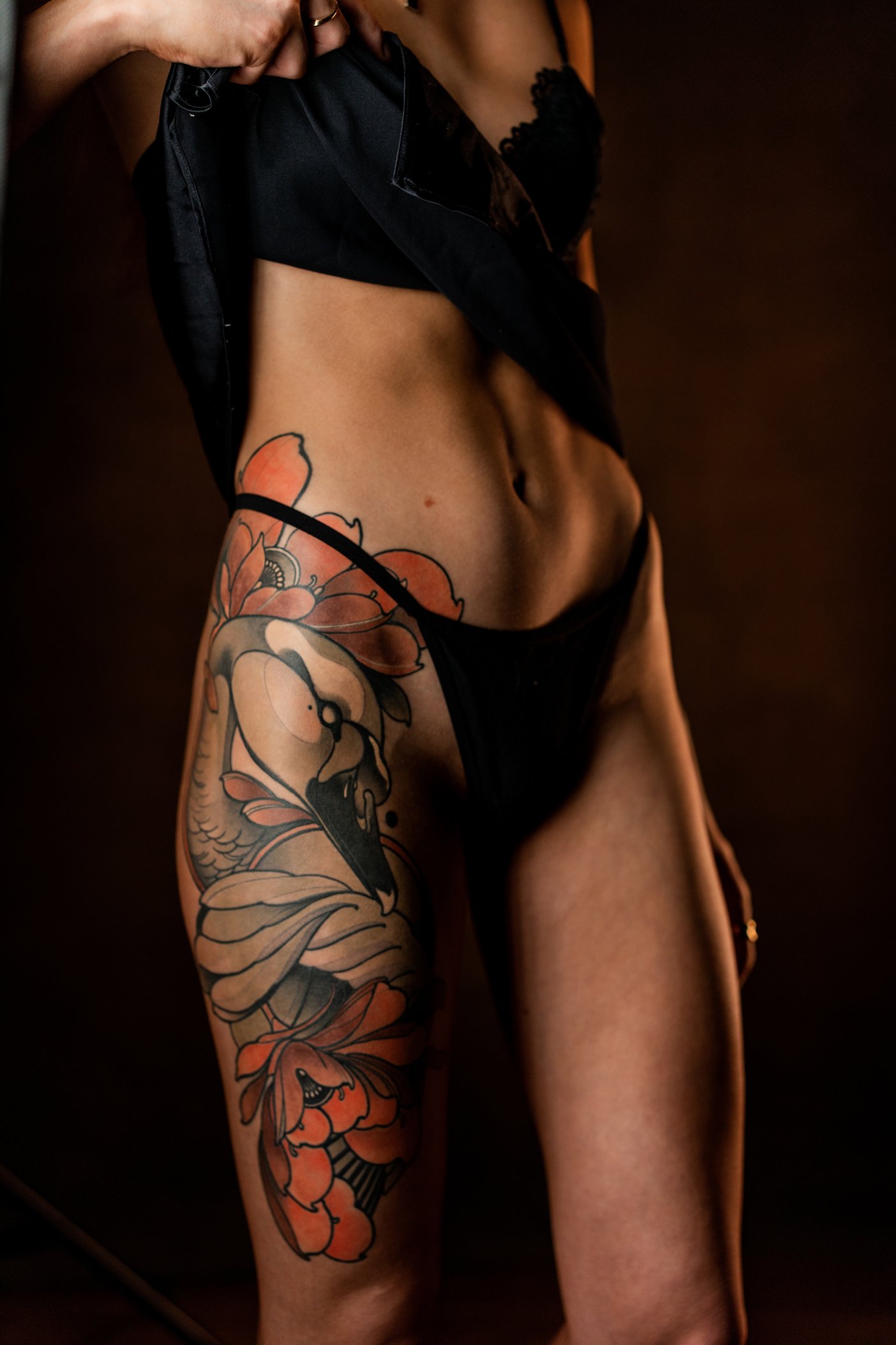 Close-up of a woman wearing black lingerie with a black shirt partially lifted, revealing a large, colorful floral tattoo on her upper thigh and hip.