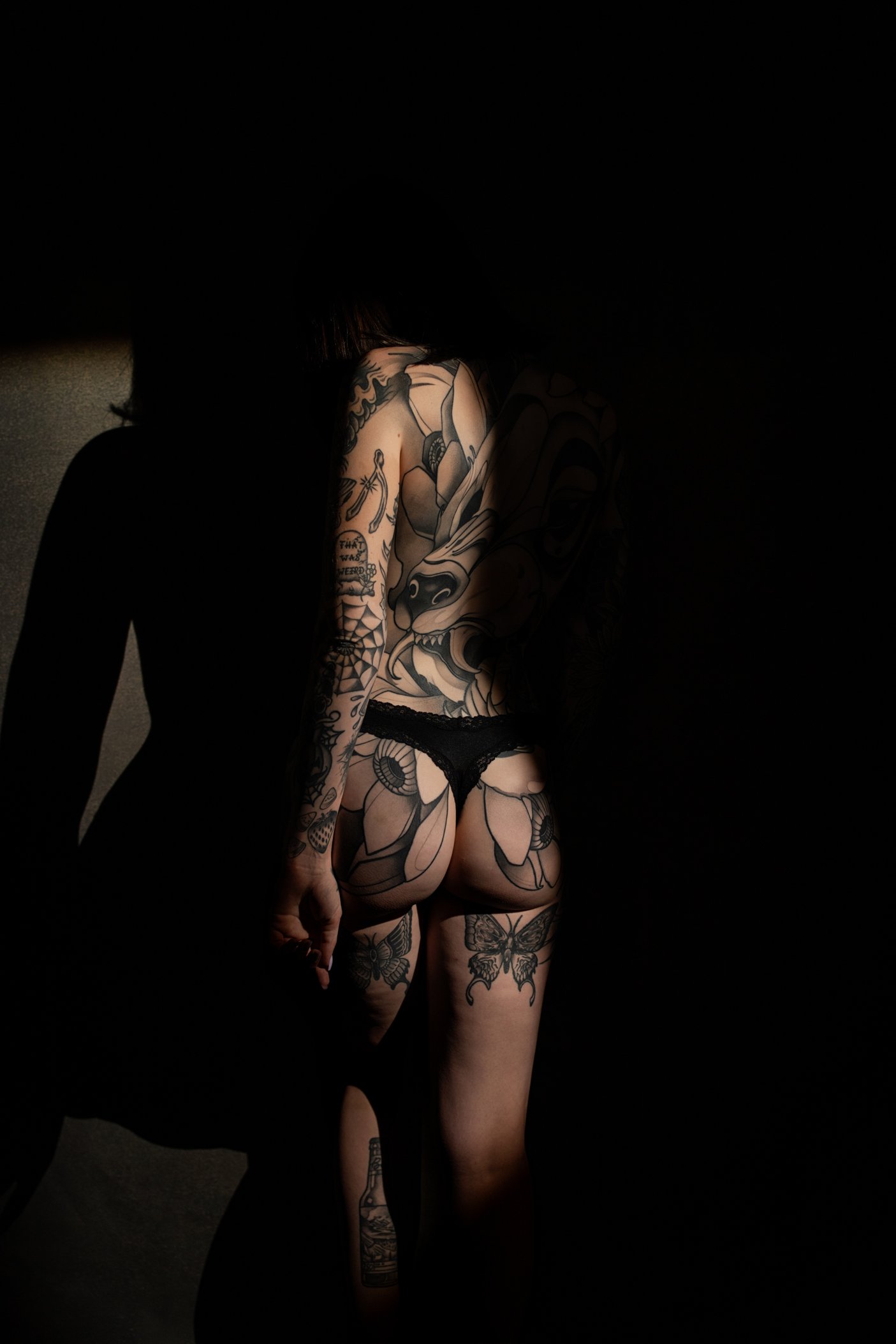 A woman with tattoos on her back and legs, wearing black lace underwear, standing against a dark background with her back facing the camera.