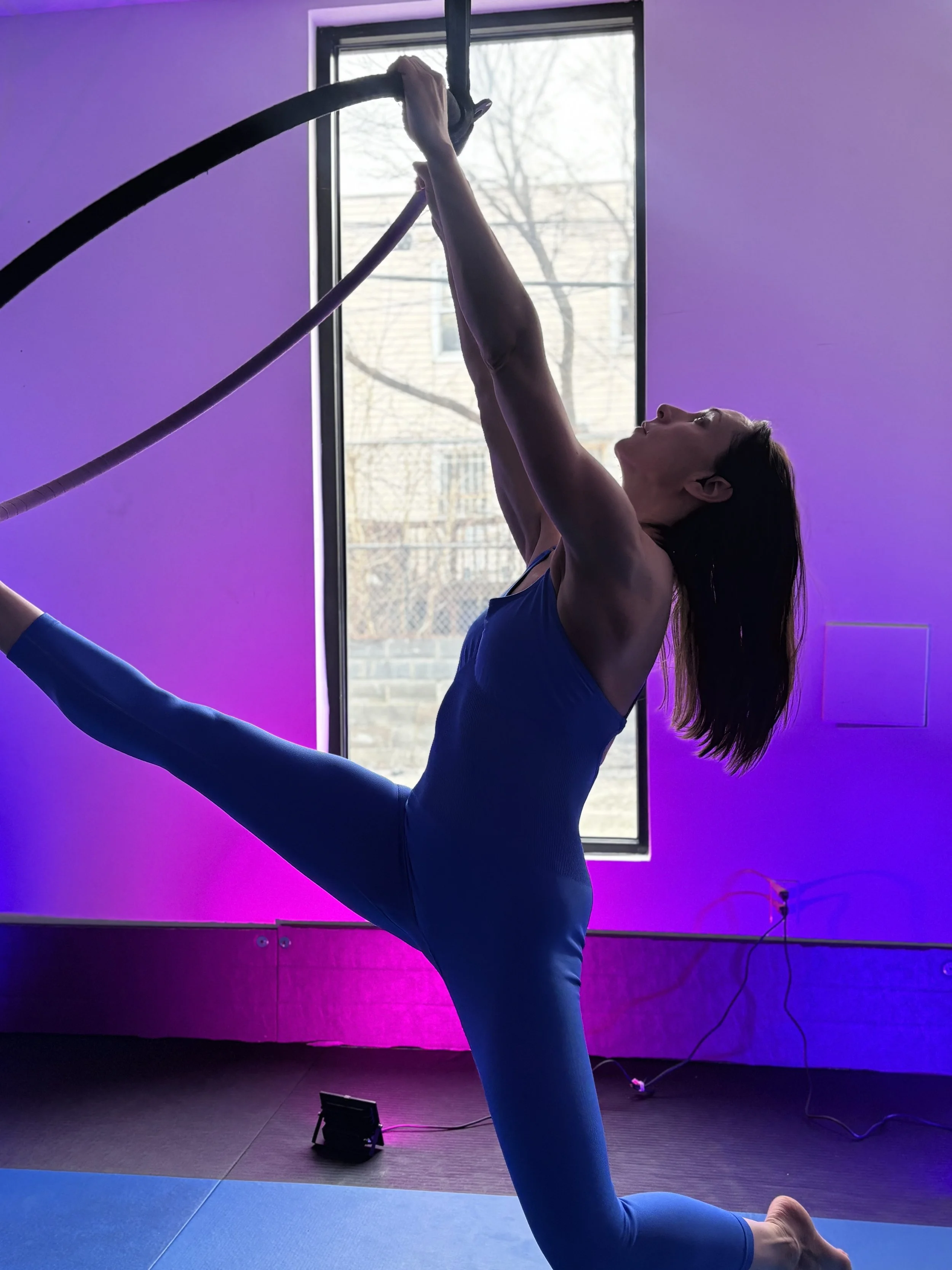 A woman practicing aerial yoga with a hoop, performing a pose in a room with purple and pink lighting, near a window showing trees outside.