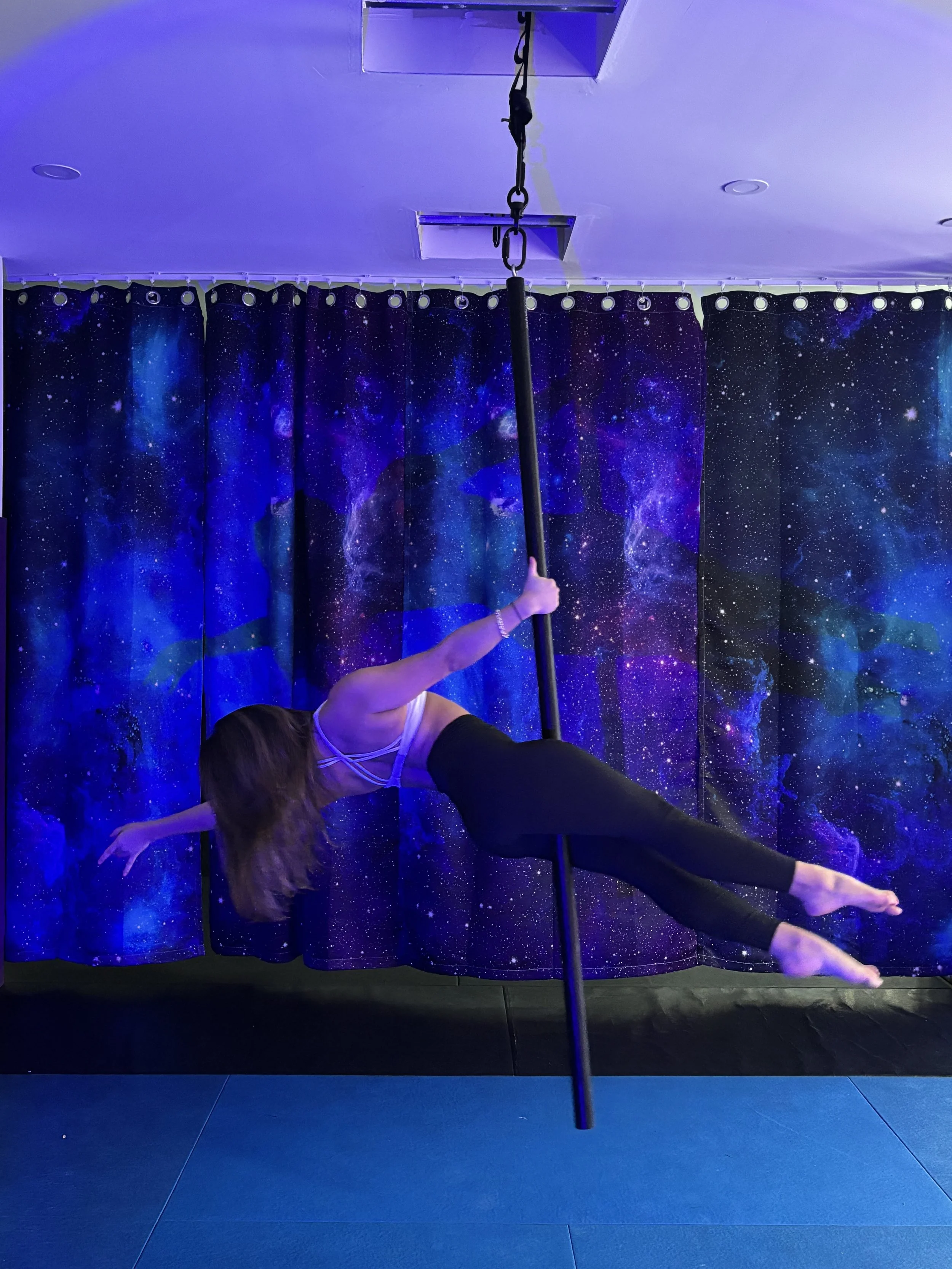 A woman practicing pole dance in front of a galaxy-themed curtain, hanging upside down on a vertical pole, in a room with blue lighting.