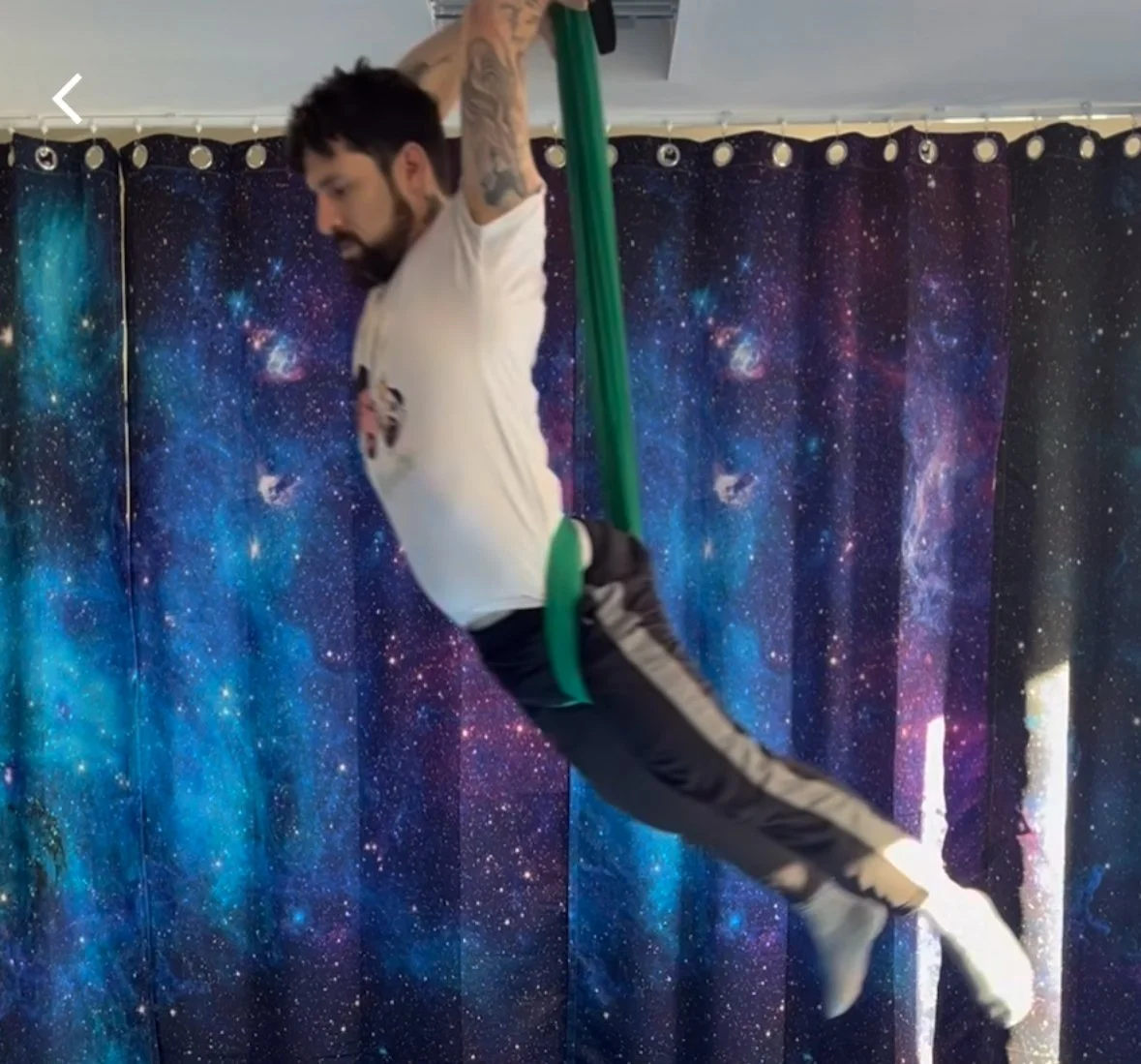A man with tattoos on his arms is performing aerial yoga or exercise using green aerial silk fabric in front of a starry galaxy curtain.
