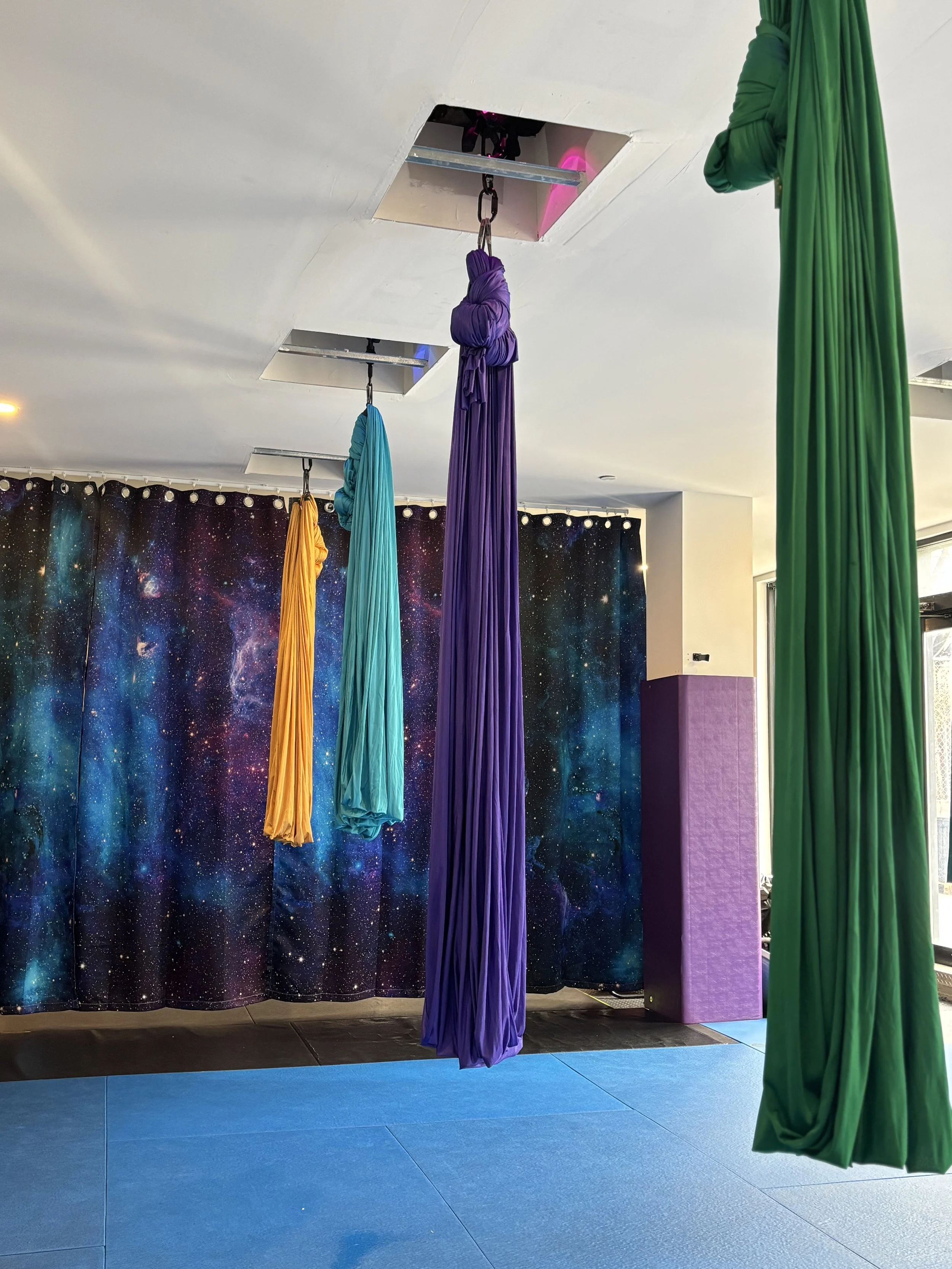 Colorful aerial silks hanging from the ceiling in a space with a galaxy-themed curtain background and blue padded flooring.
