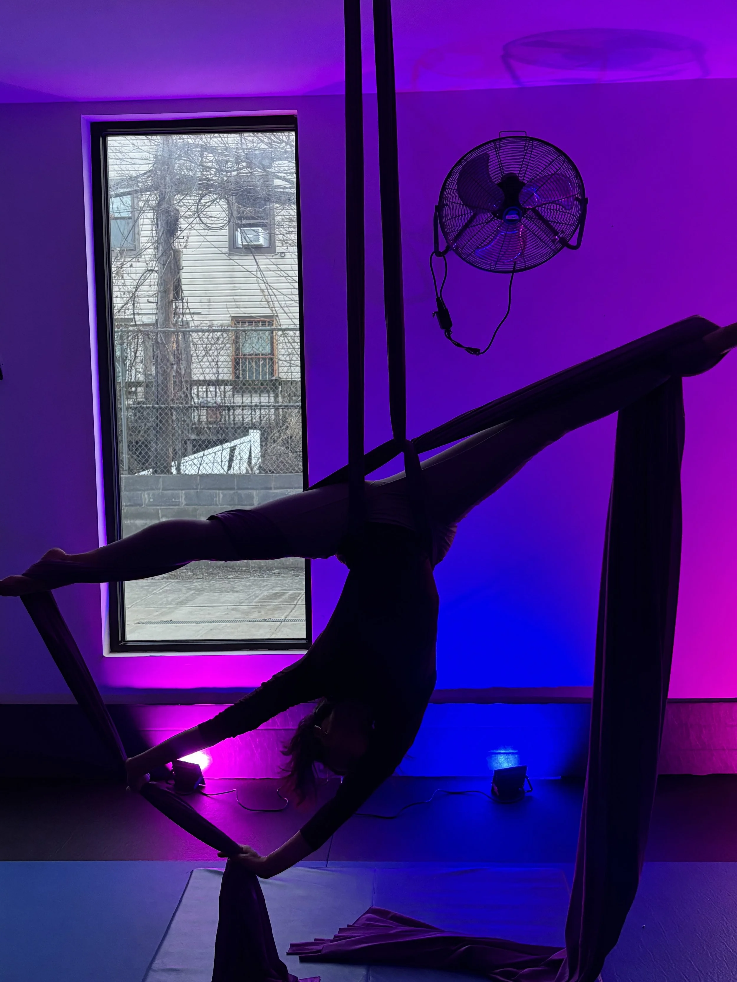 Silhouette of a person performing an aerial silk yoga pose indoors, with purple and blue ambient lighting, a large window showing an outdoor scene, a wall fan, and yoga equipment on the floor.