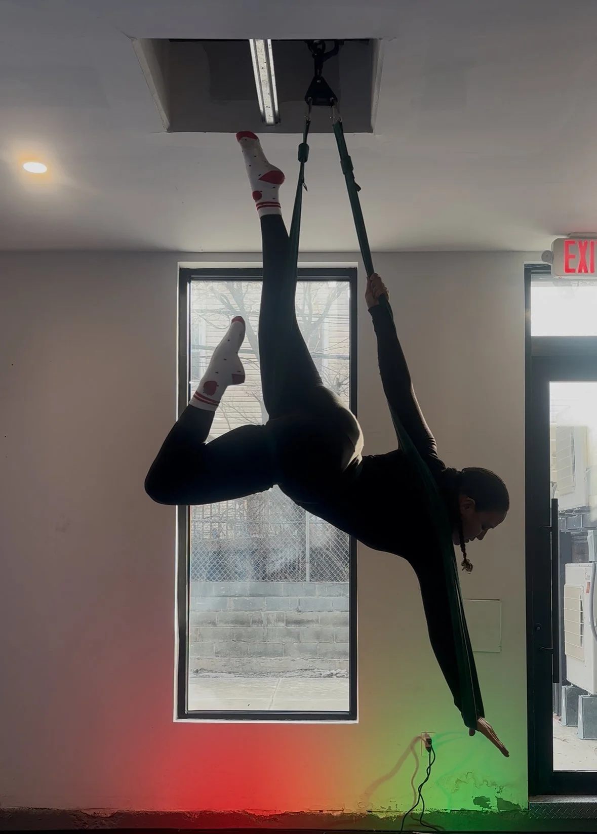 A person practicing aerial silk performance upside down, hanging from a strap attached to a ceiling open hatch, inside a room with windows and an exit sign.