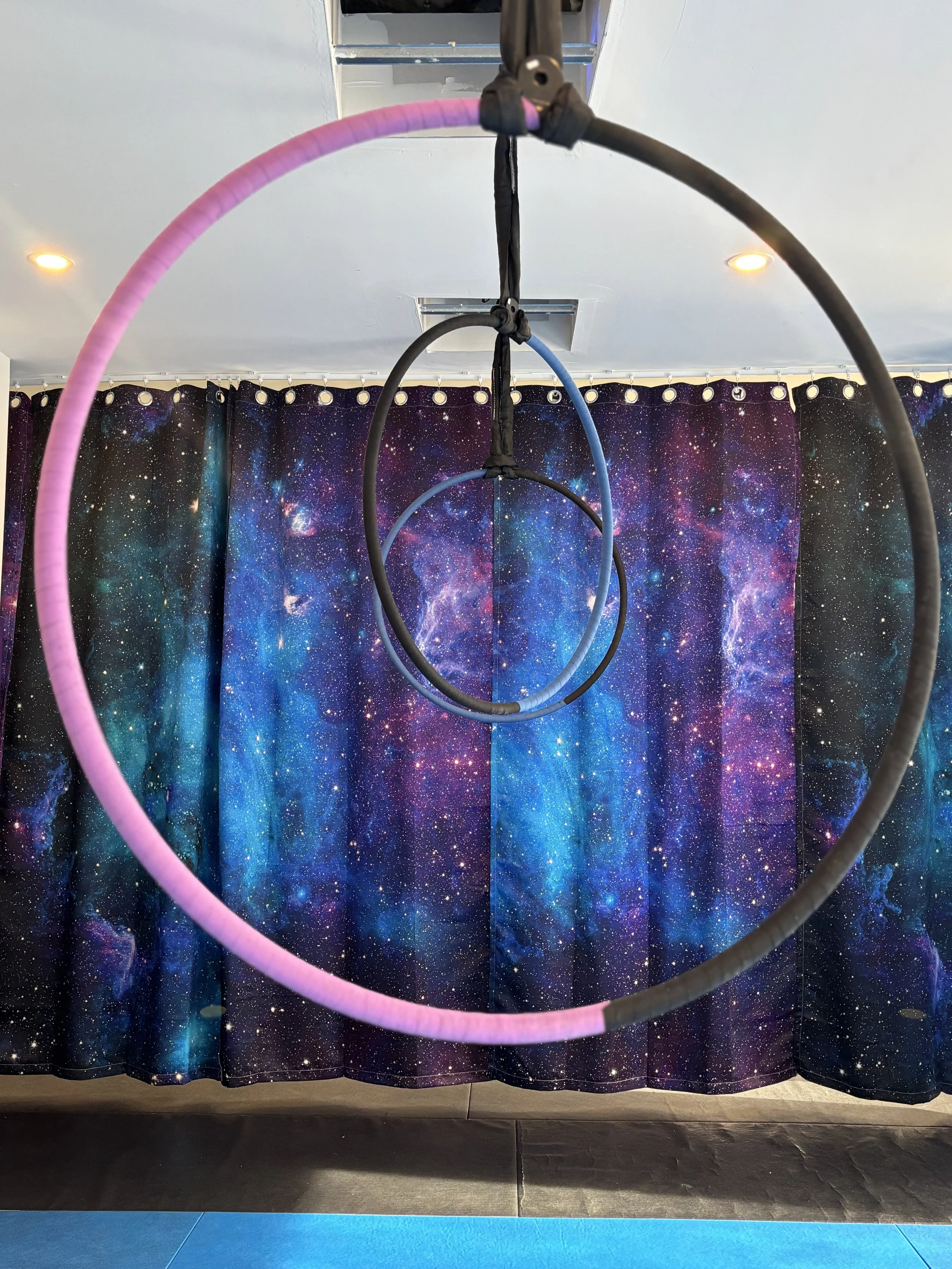 Three aerial hoops hanging from the ceiling in front of a galaxy-themed curtain.