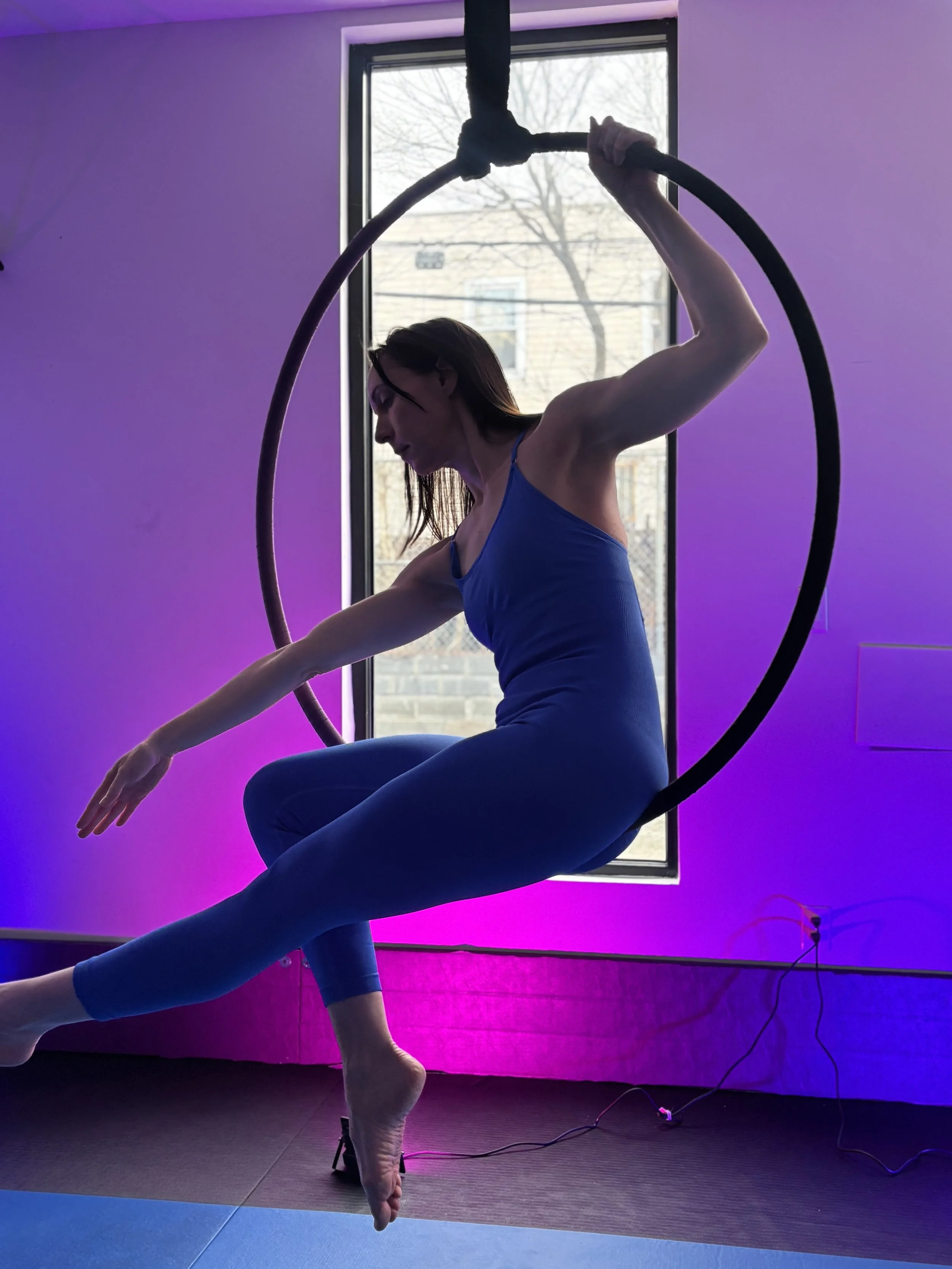 A woman performing an aerial silk pose inside a room with a large window and colorful LED lighting.