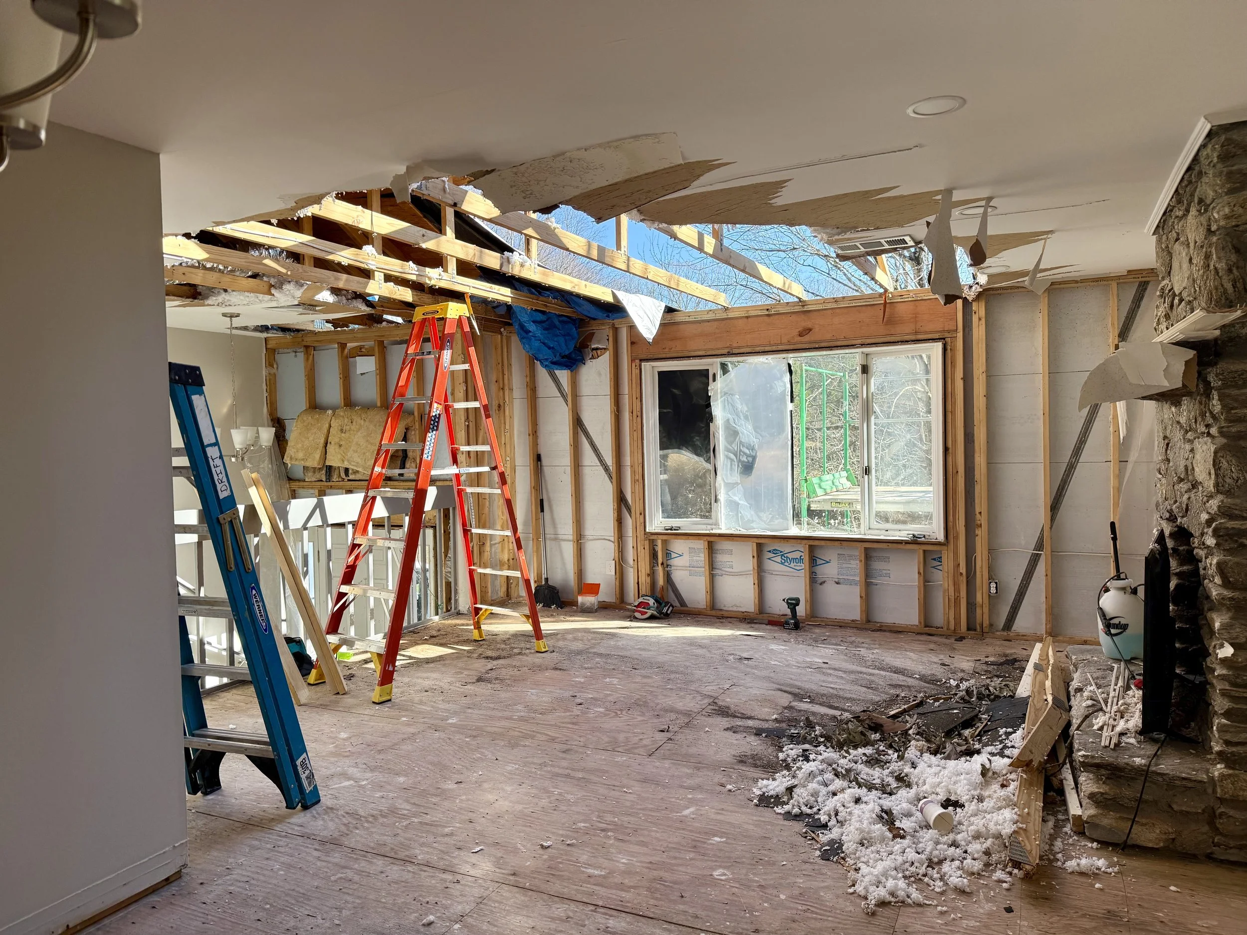 Interior of a house under renovation showing exposed wooden framing, tear-down of a ceiling, a window with outdoor view, ladders, construction debris, and snow on the floor.