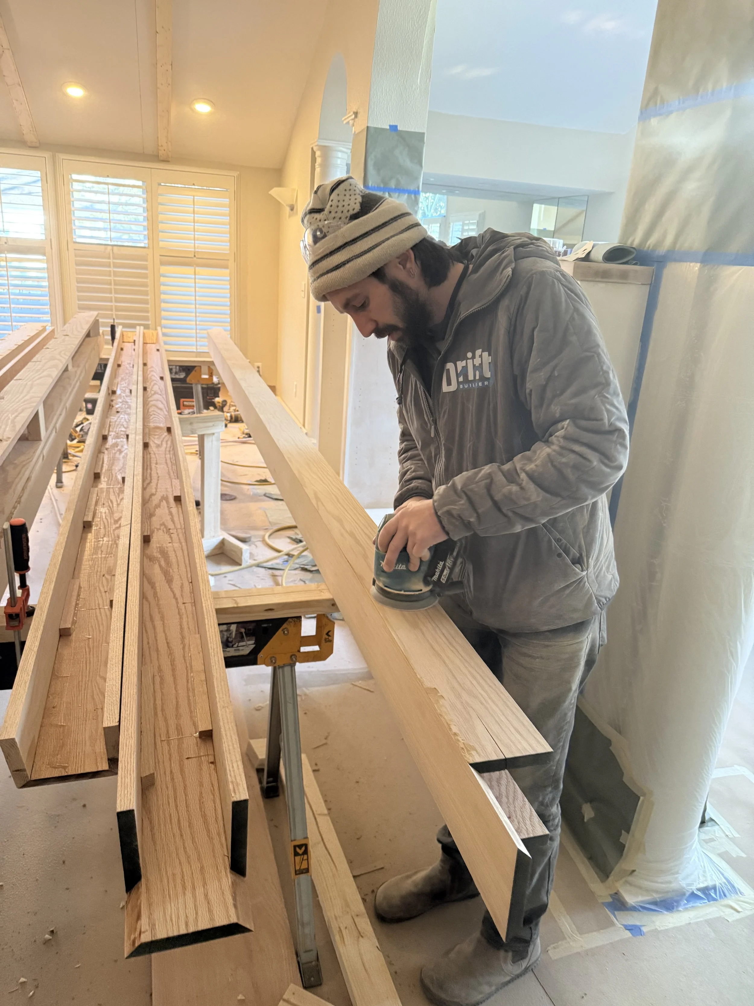A man working on woodworking project, sanding a piece of wood in a room with large windows, some covered with tape, and construction tools around.