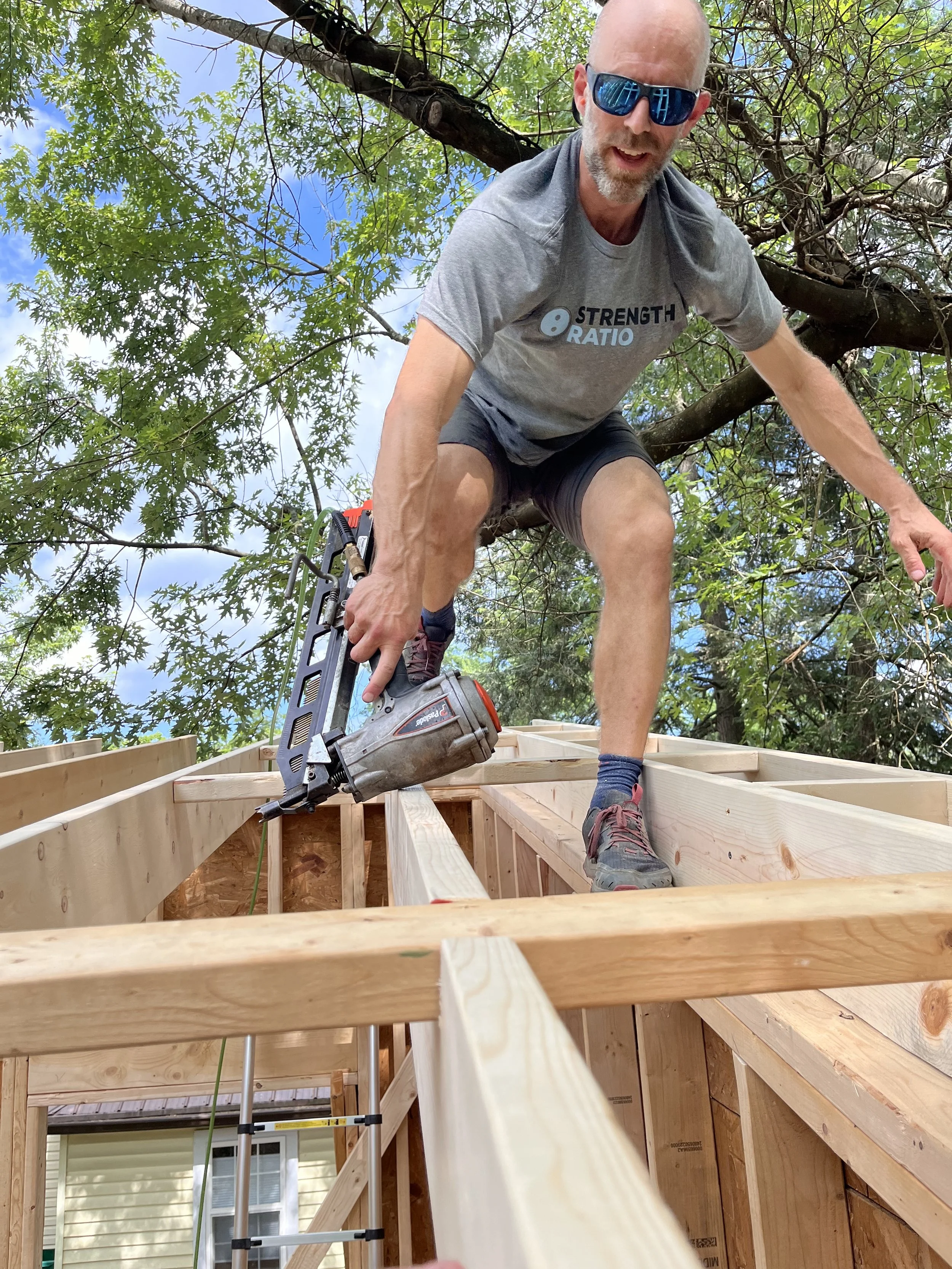 A man wearing sunglasses, a grey t-shirt with the words 'Strength Ratio,' and shorts is working on a wooden construction project outdoors, climbing on the framework of a house surrounded by green trees.
