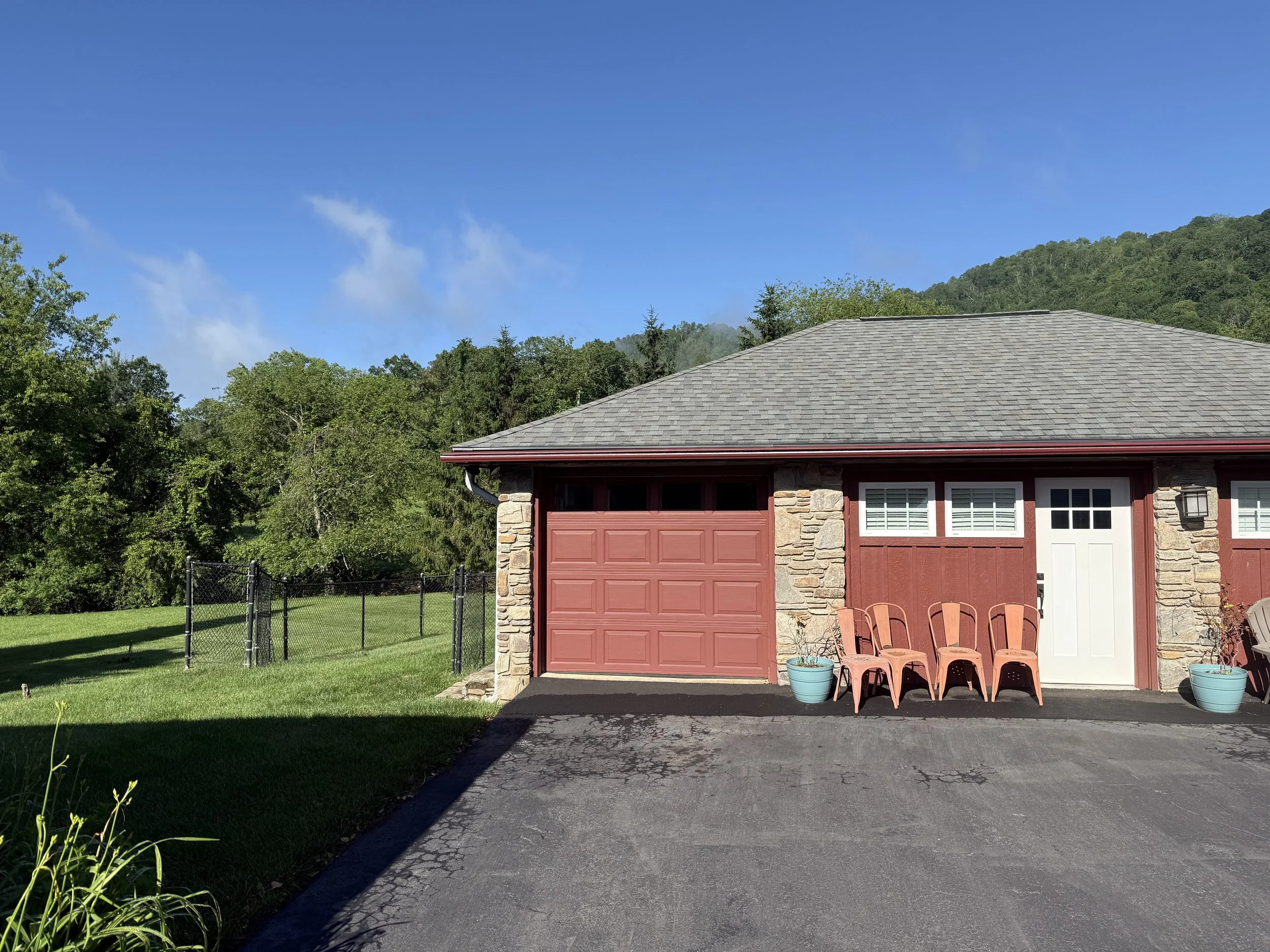 Front view of a house with a gray shingle roof, red garage door, and beige stone and siding exterior. There are four pink chairs and two blue planters outside, and a lush green lawn with trees in the background. Clear blue sky overhead.