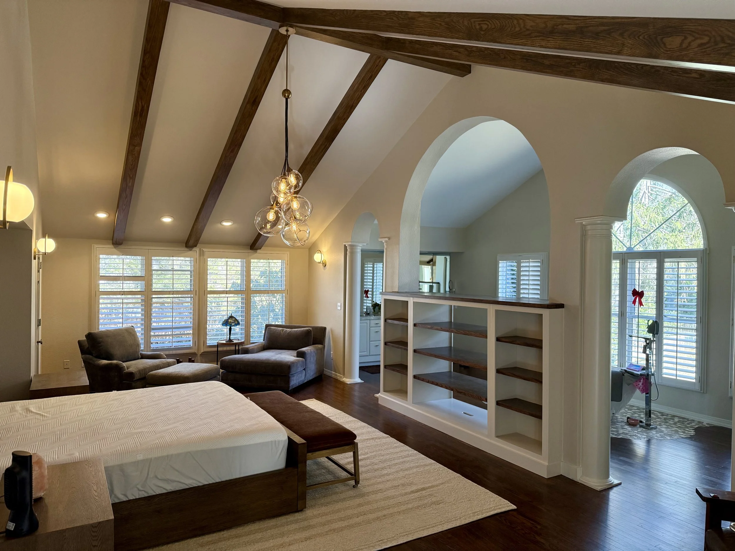 A living room with wooden beams on the ceiling, a chandelier with multiple bulbs, large windows with white shutters, a beige armchair, a lamp, and a white shelving unit near a bay window decorated with red bows.