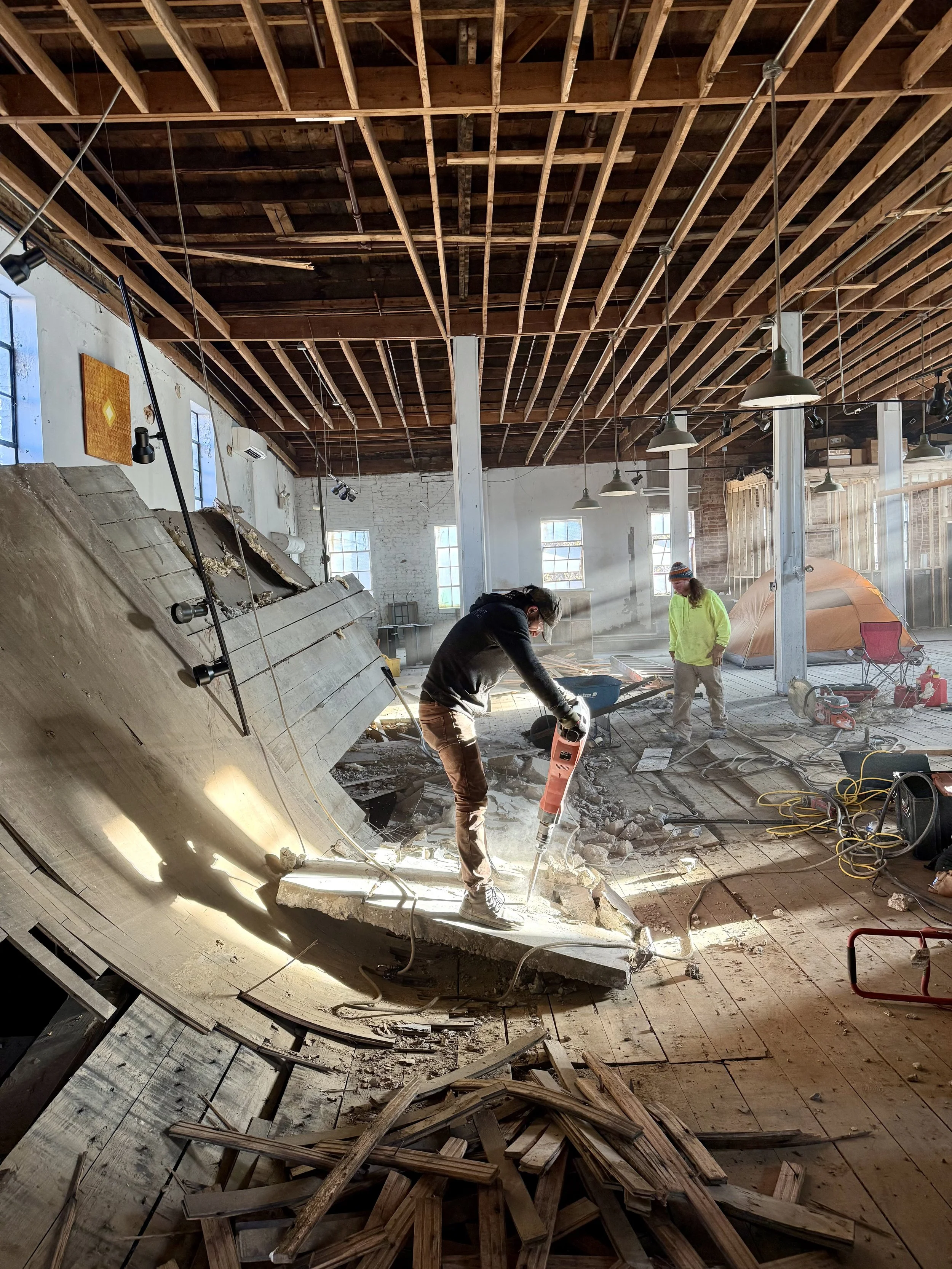 Construction workers demolishing a wall in an indoor building with exposed ceiling beams, large windows, and construction tools scattered on the floor.