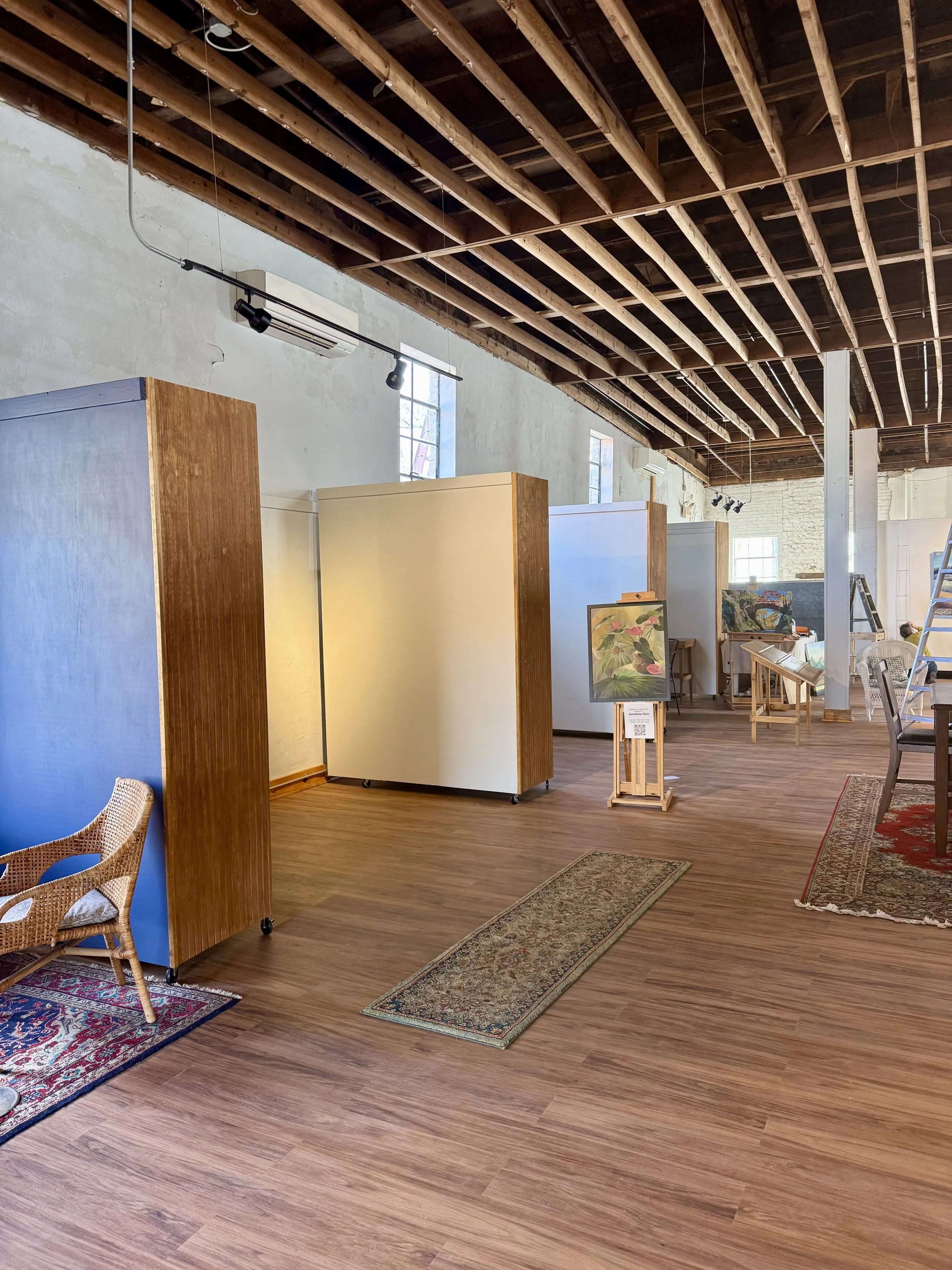 An indoor space with wooden ceiling beams, along with exhibition booths and artwork on display, including framed paintings and rugs on the floor.