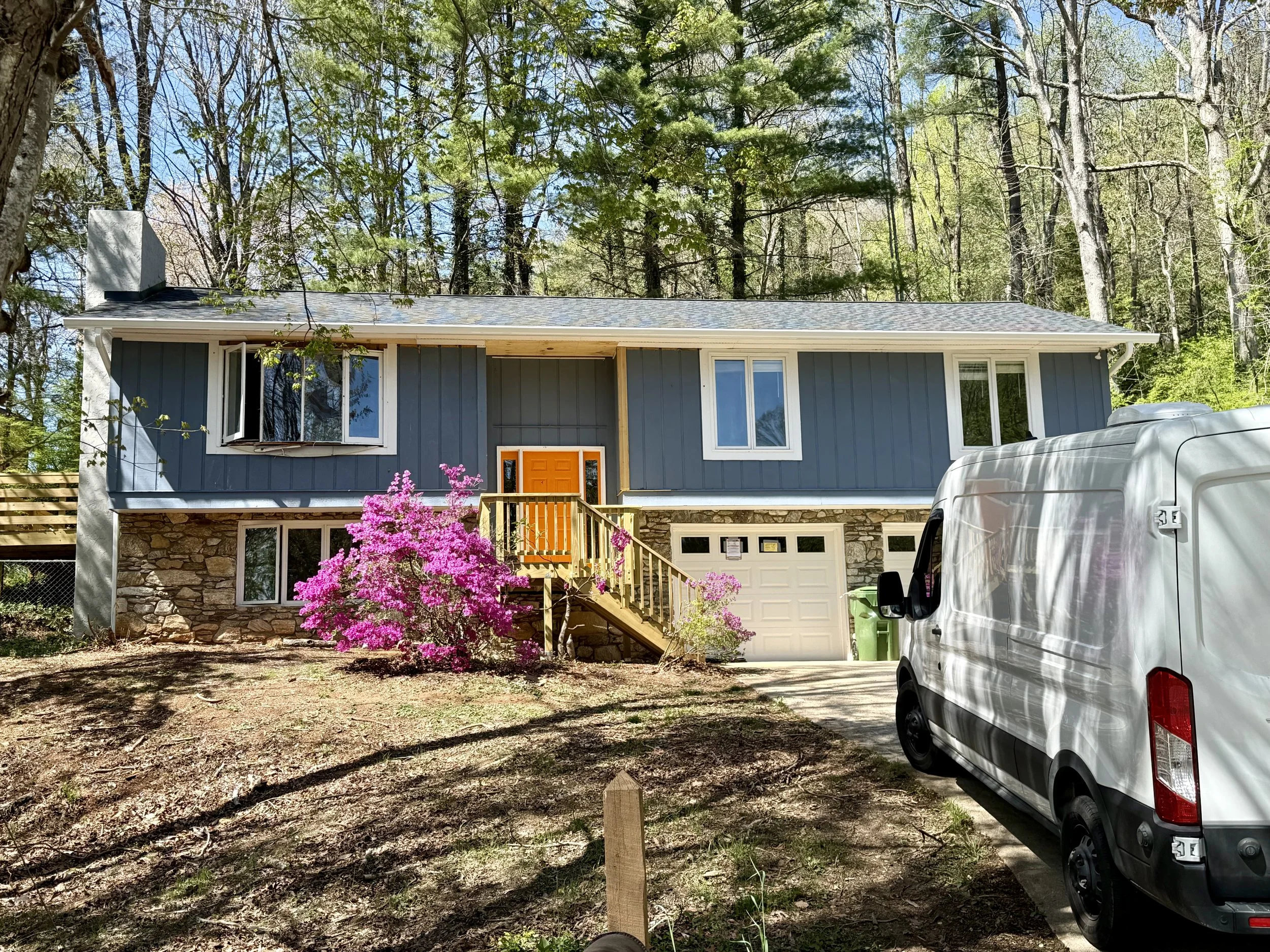 A two-story house with a blue exterior and a gray roof, surrounded by trees and a driveway with a white van parked on the right.