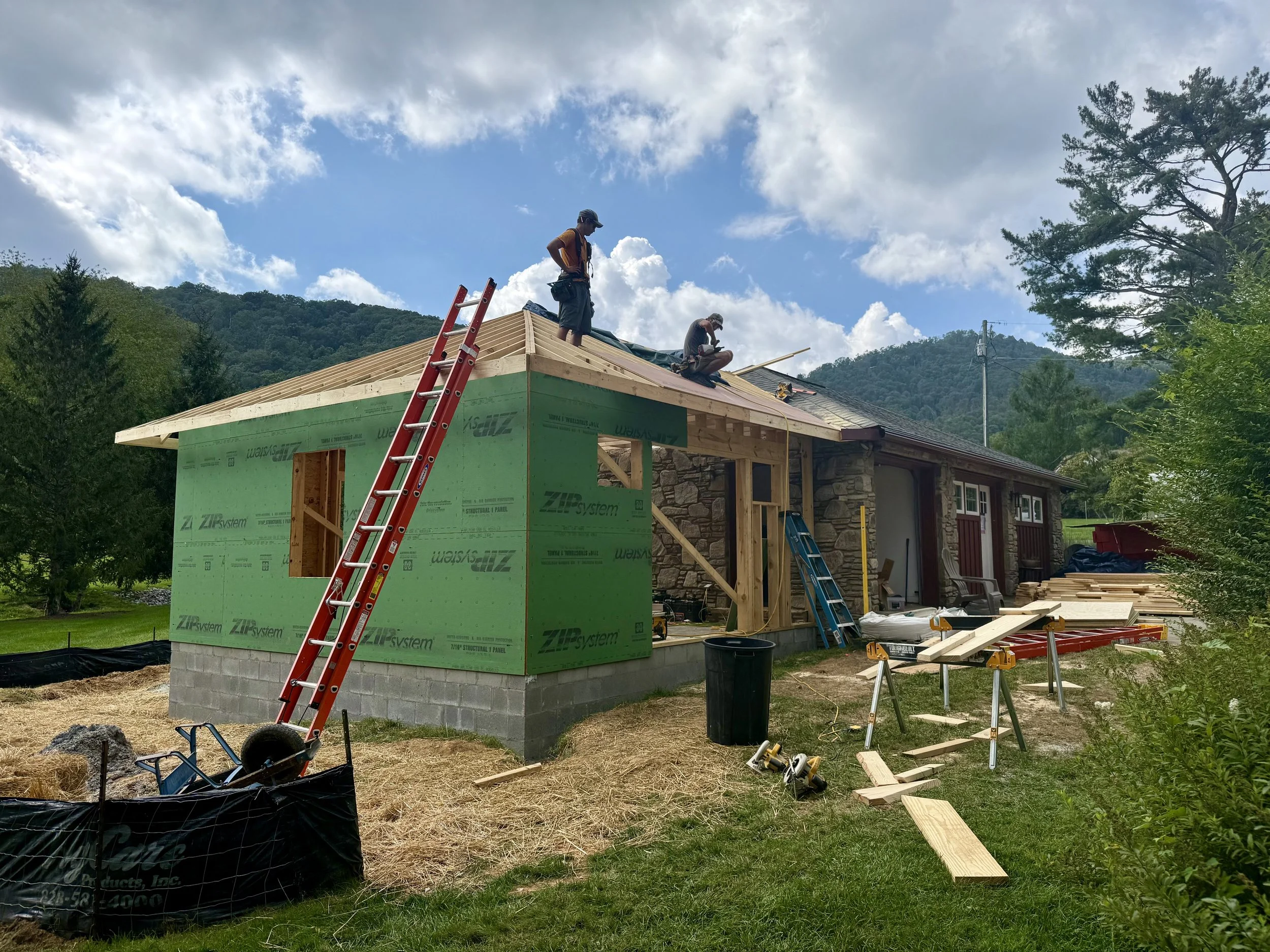Workers building a wooden house on a sunny day with a blue sky and scattered clouds, surrounded by trees and mountains.