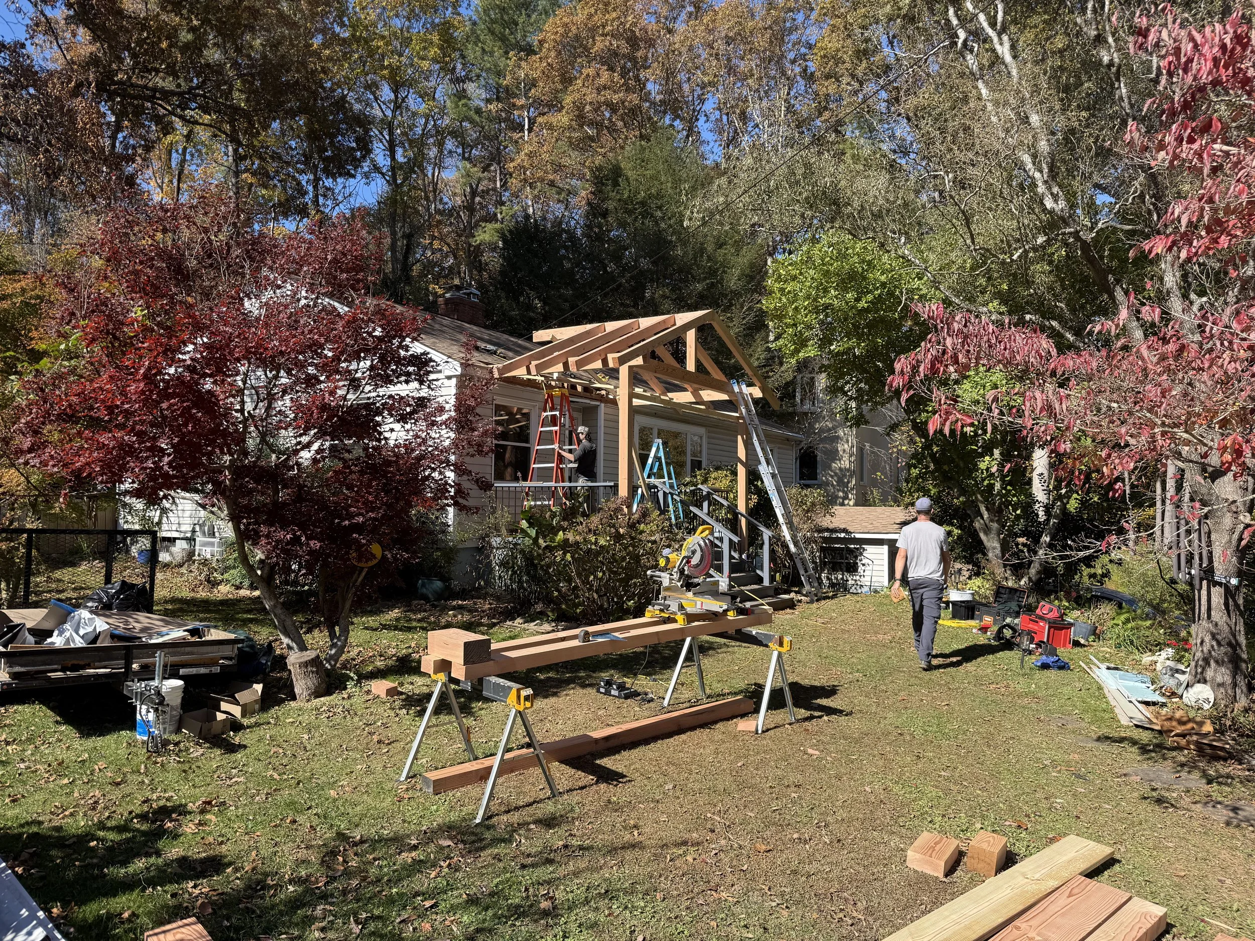 A backyard with trees and a house under construction or renovation. Several ladders, tools, and wood pieces are scattered around as workers build a wooden structure attached to the house.