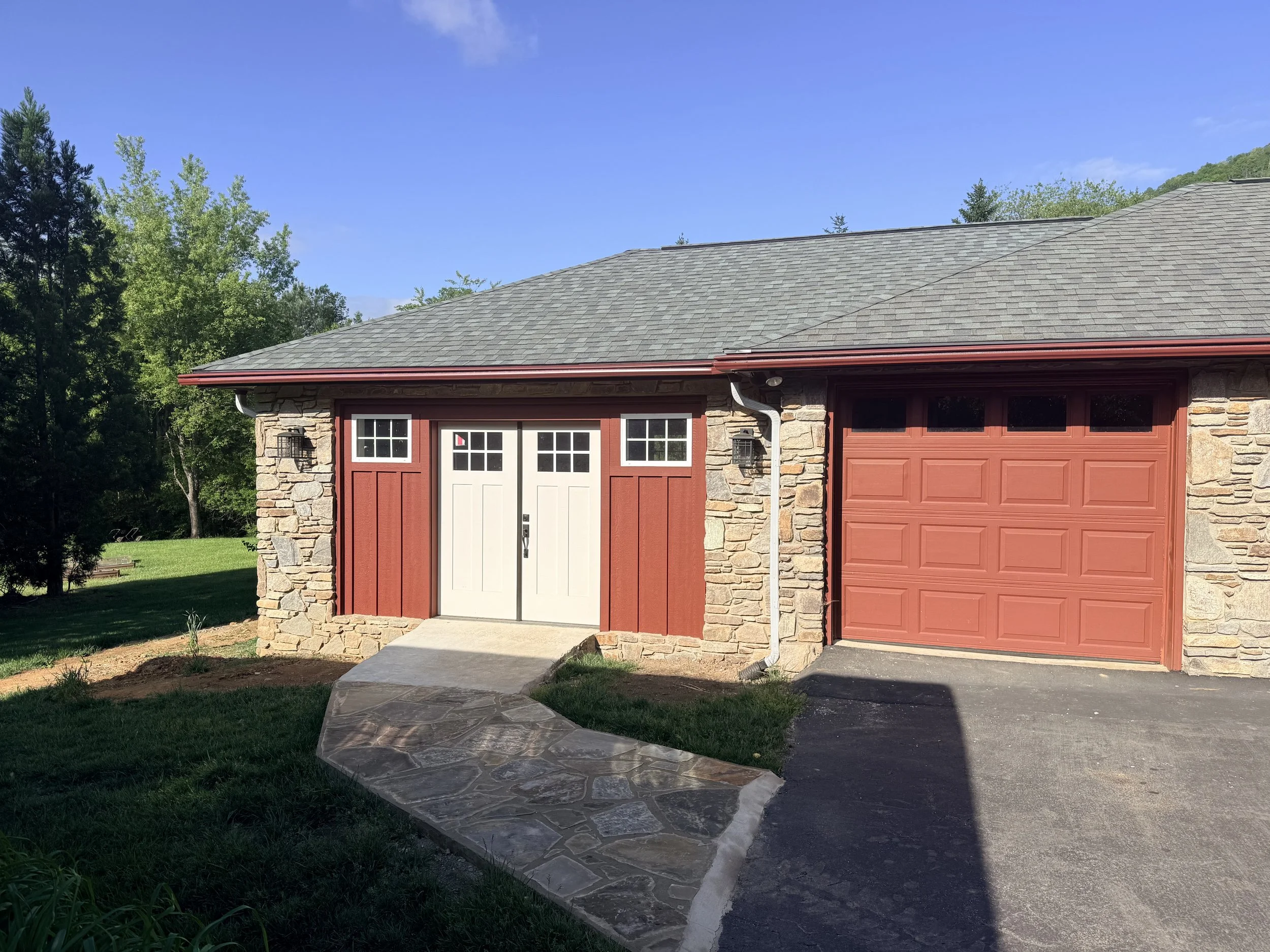A house with a stone and red exterior, a gray shingle roof, and a red garage door, with a walkway leading to the front and surrounded by trees and greenery under a clear blue sky.
