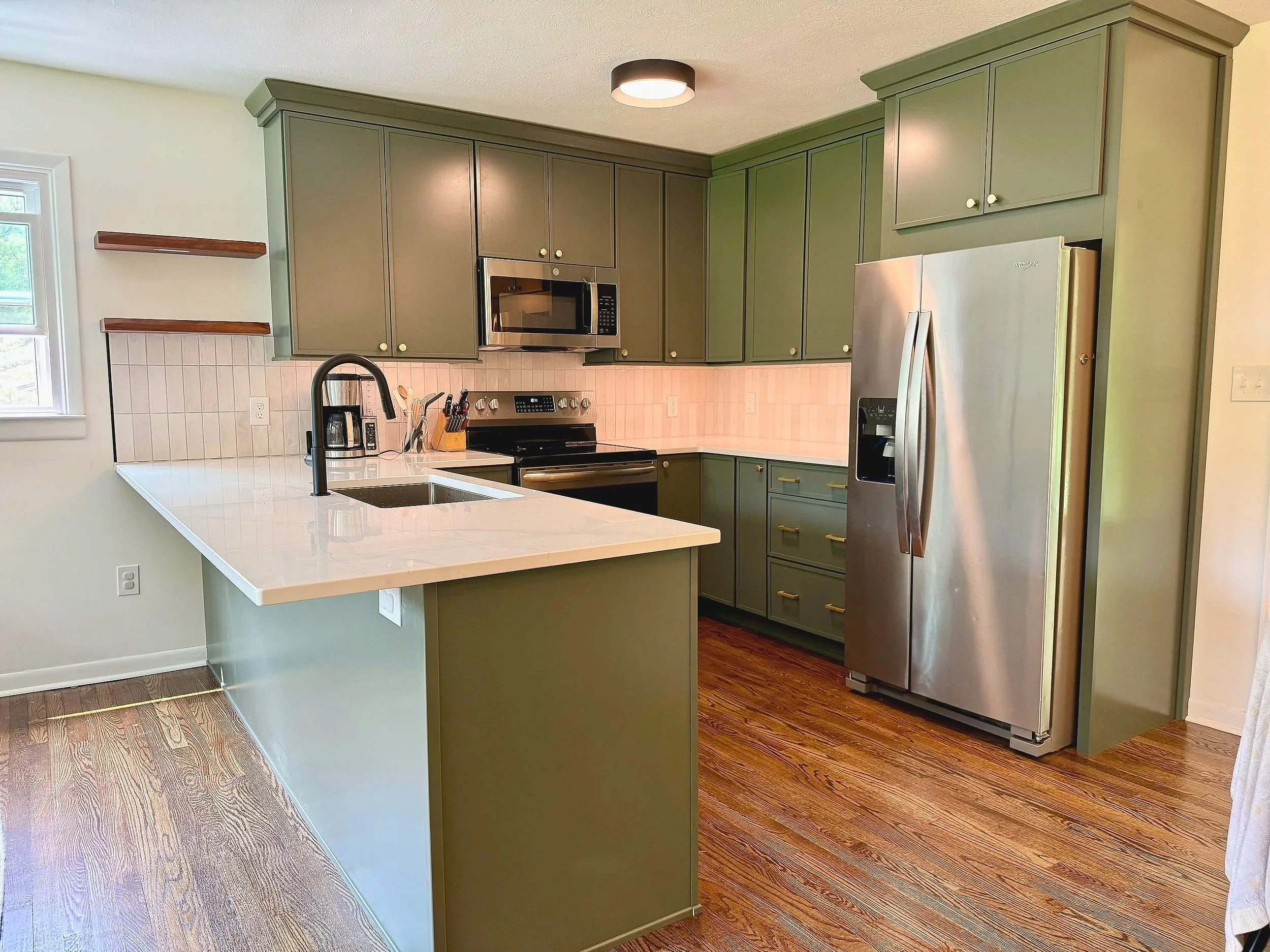 Modern kitchen with green cabinetry, stainless steel appliances, a white countertop island with a sink and black faucet, hardwood floors, a window, and small wooden shelf accents.