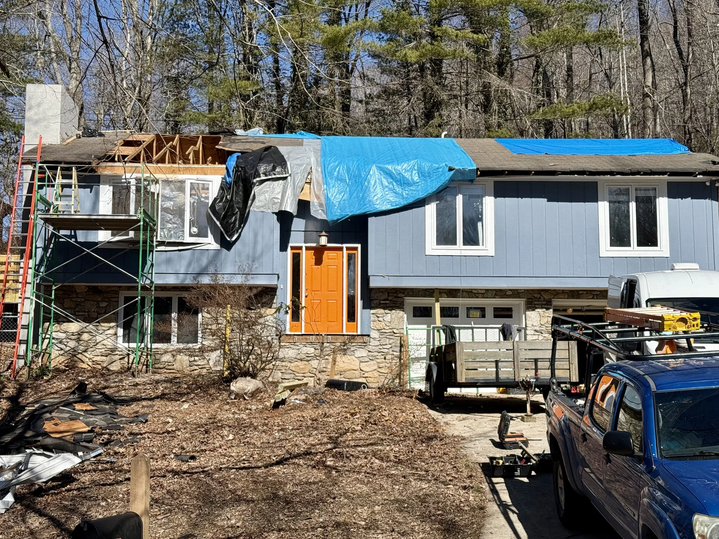 A house undergoing roof renovation with partial roof removal, construction scaffolding, and materials covered with blue and black tarps. The house is blue with an orange front door and white window frames, and a wooded area is in the background.