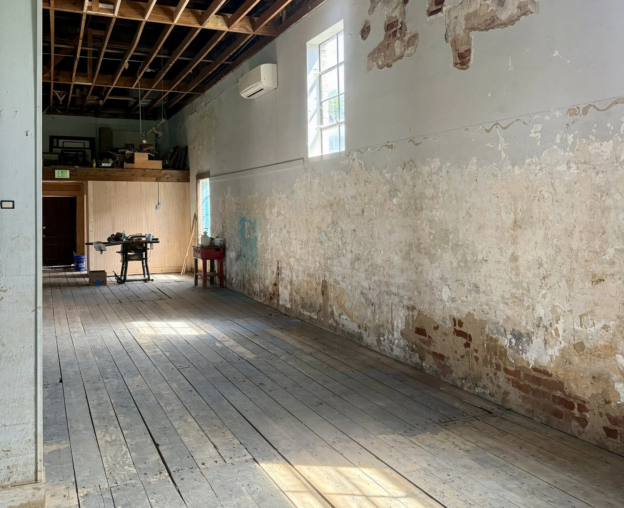 Interior of a room under renovation with exposed brick and plastered walls, unfinished wooden floors, a large window, an air conditioner, and construction tools and materials.