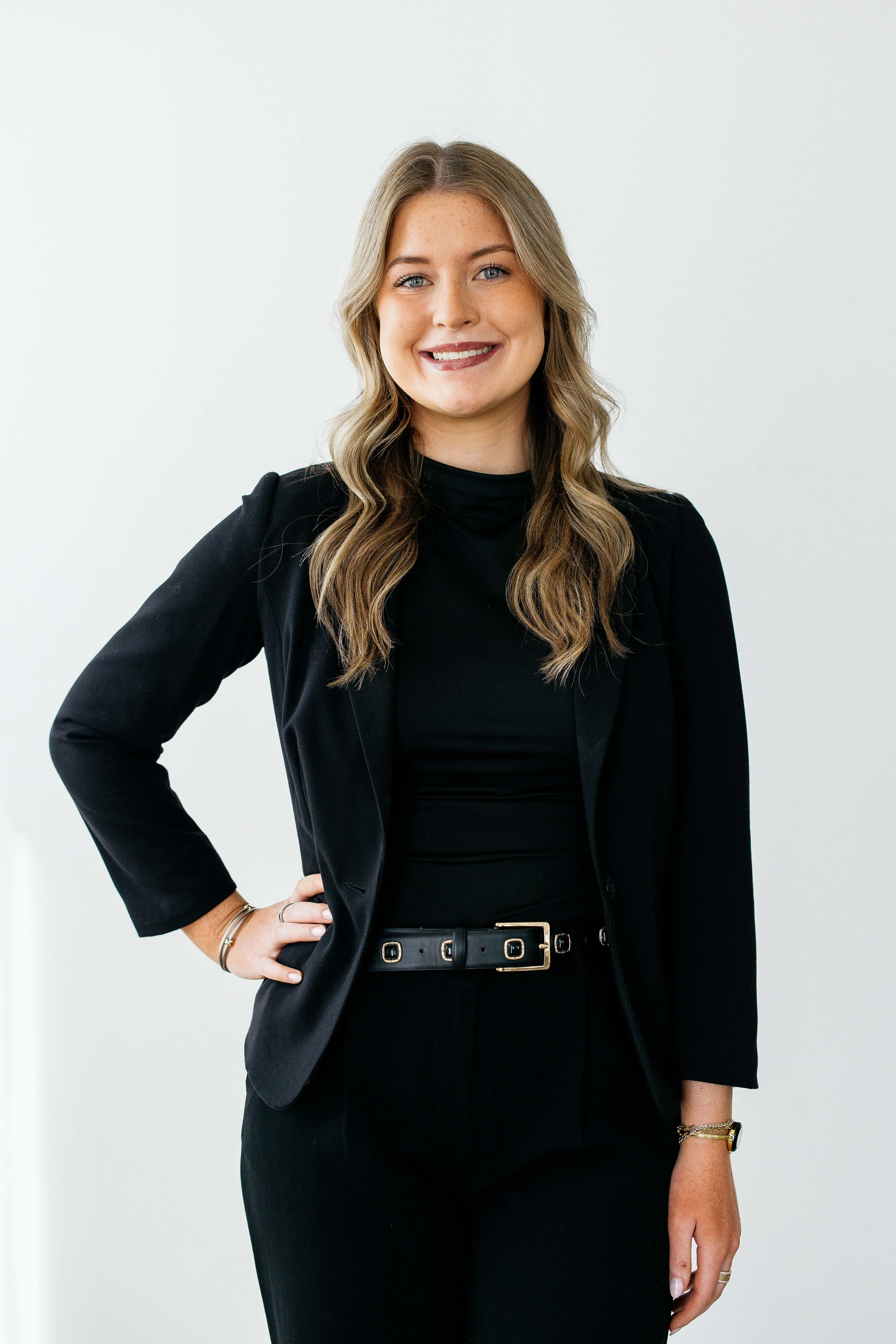 A young woman sitting on a wooden stool, wearing a black sleeveless turtleneck dress and black high heels, smiling at the camera against a plain white background.